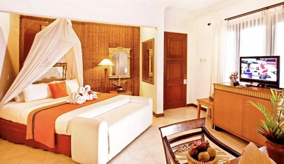 A hotel room with a large bed adorned with towel swans, a wooden cabinet with a television, and a sitting area near a window with sheer curtains.