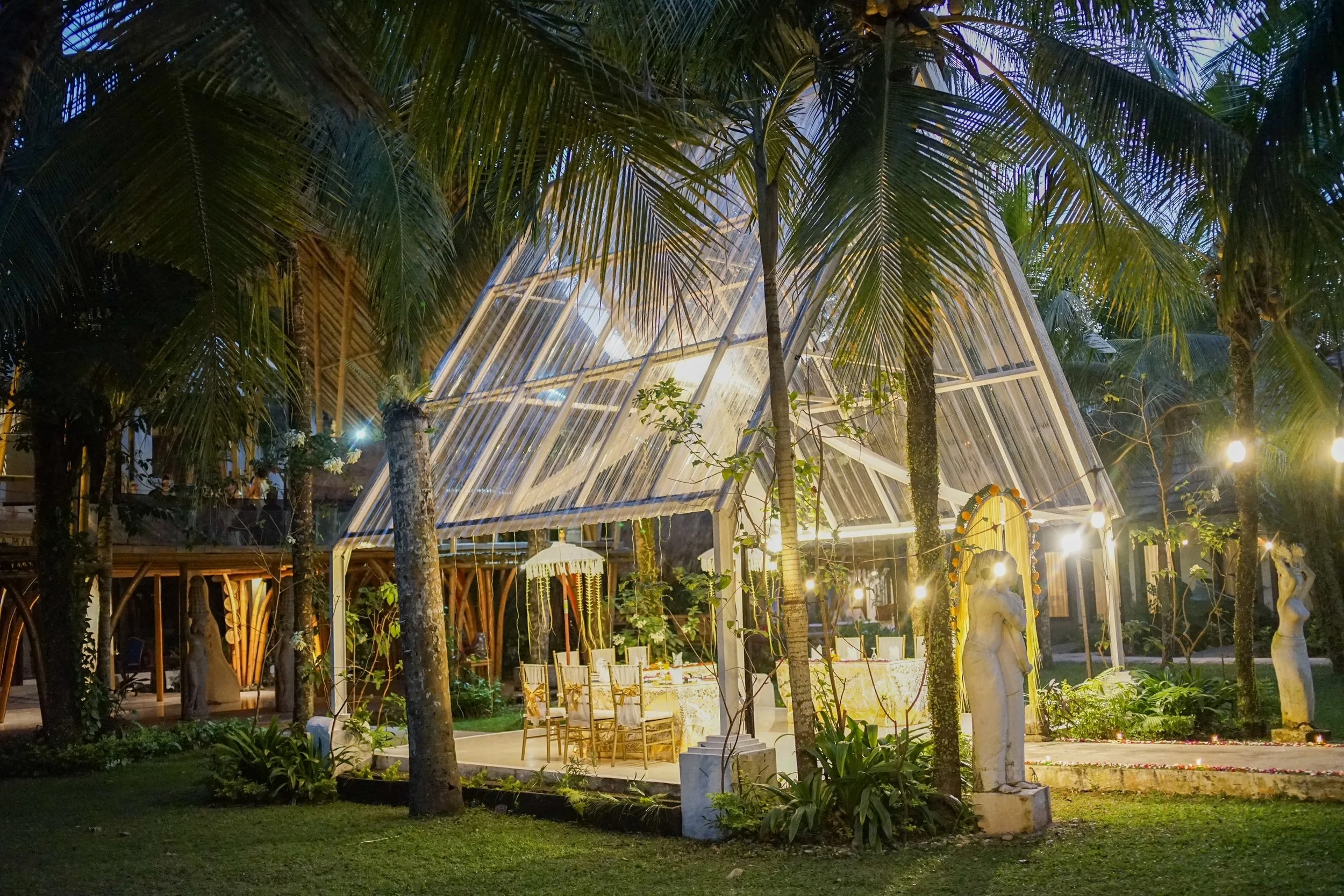 Nighttime outdoor scene with illuminated modern glass structure surrounded by palm trees and garden statues.
