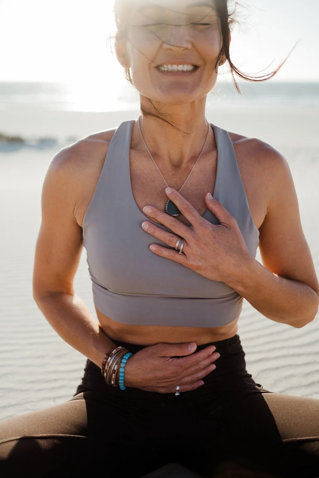 A woman with eyes closed smiling while practicing yoga outdoors on a beach, wearing a gray sports bra and black leggings, with jewelry and her hands resting on her chest and stomach.
