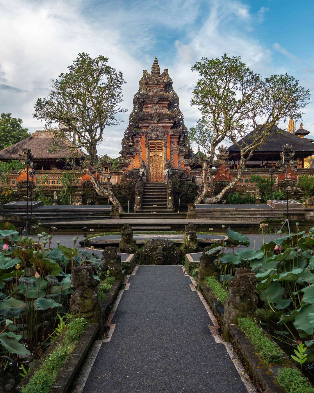 An ornate Balinese temple with traditional architecture, stone stairs, and lush greenery, including lotus plants with pink flowers in the foreground, under a partly cloudy sky.