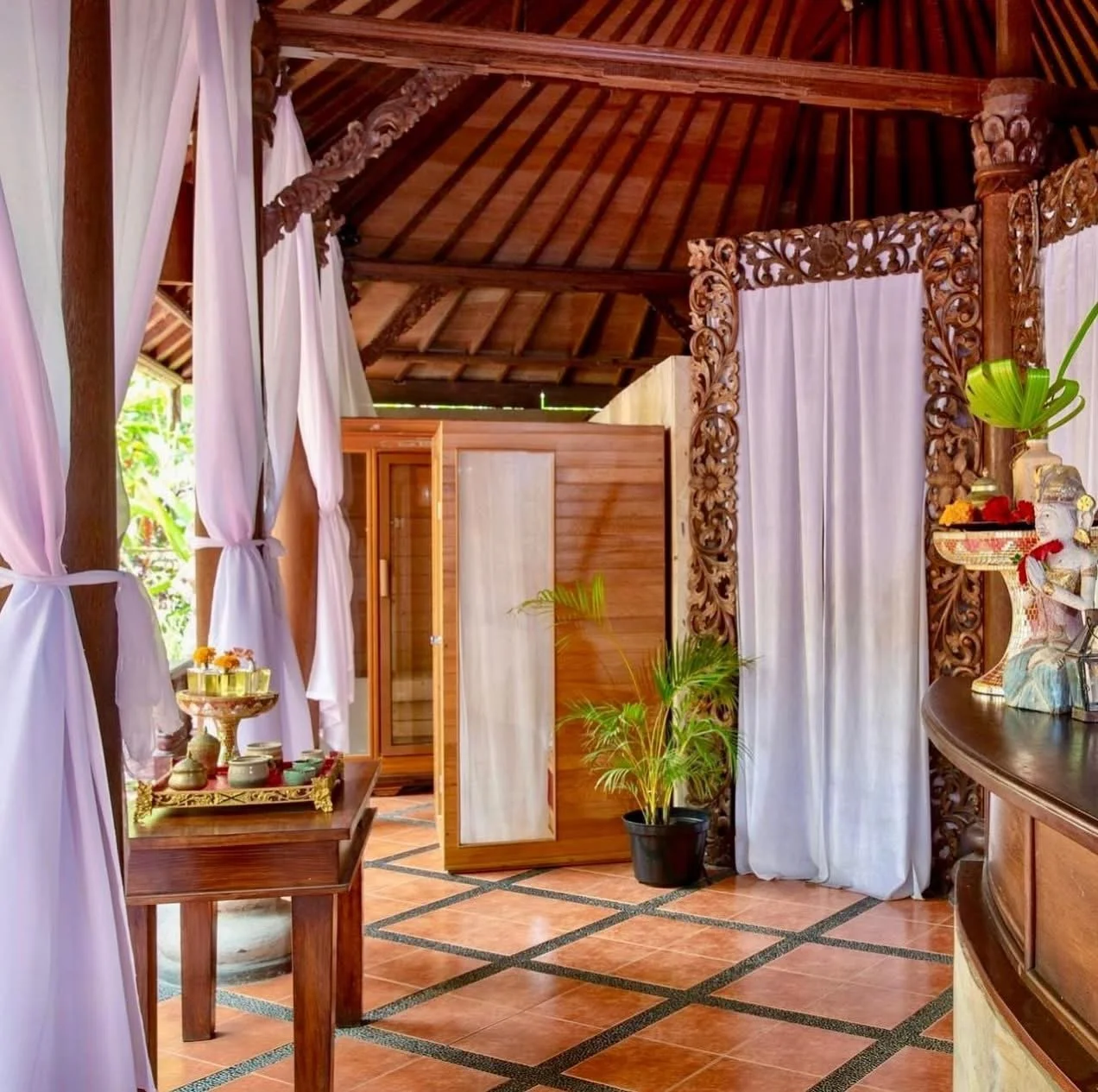 Interior of a tropical-style room with wooden furniture, white curtains, and decorative carvings. There is a wooden table with a tray of items, some potted plants, and a small wooden enclosure or shower stall.