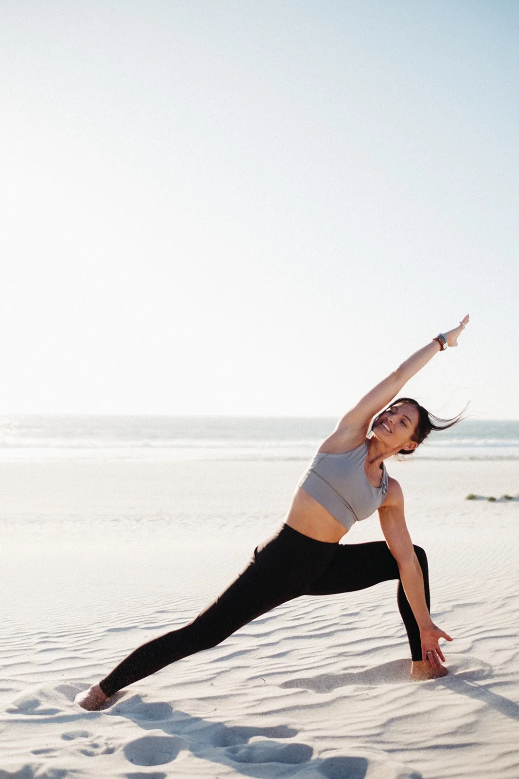 A woman practicing yoga on the beach, performing a side stretch pose with one arm extended upwards and the other hand on the sand, with the ocean and sky in the background.