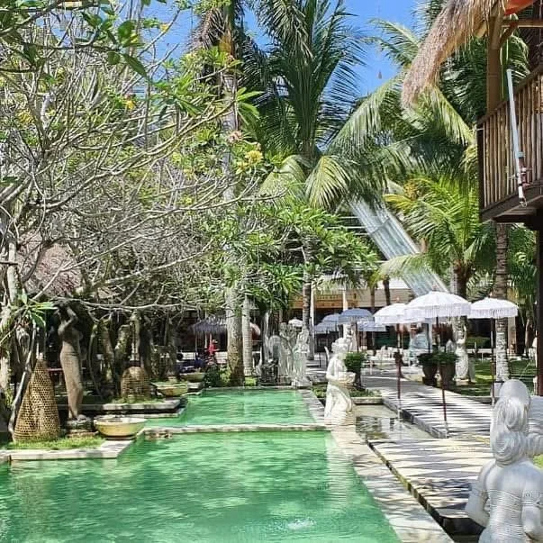 A peaceful outdoor scene with a narrow pool of water surrounded by statues, trees, and umbrellas in a tropical setting.