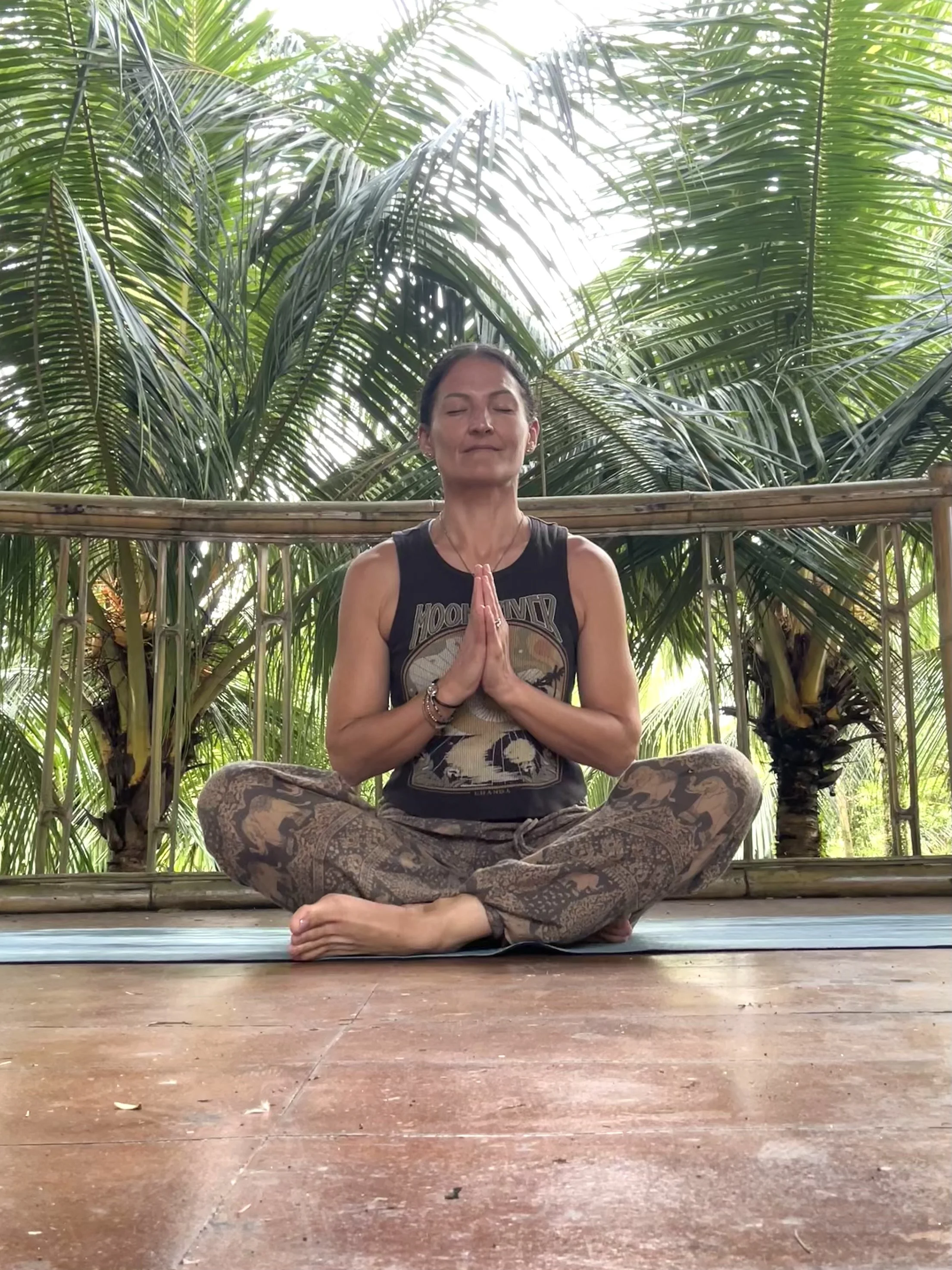 A woman practicing yoga outdoors, seated cross-legged with hands together in prayer position, surrounded by lush green palm trees.