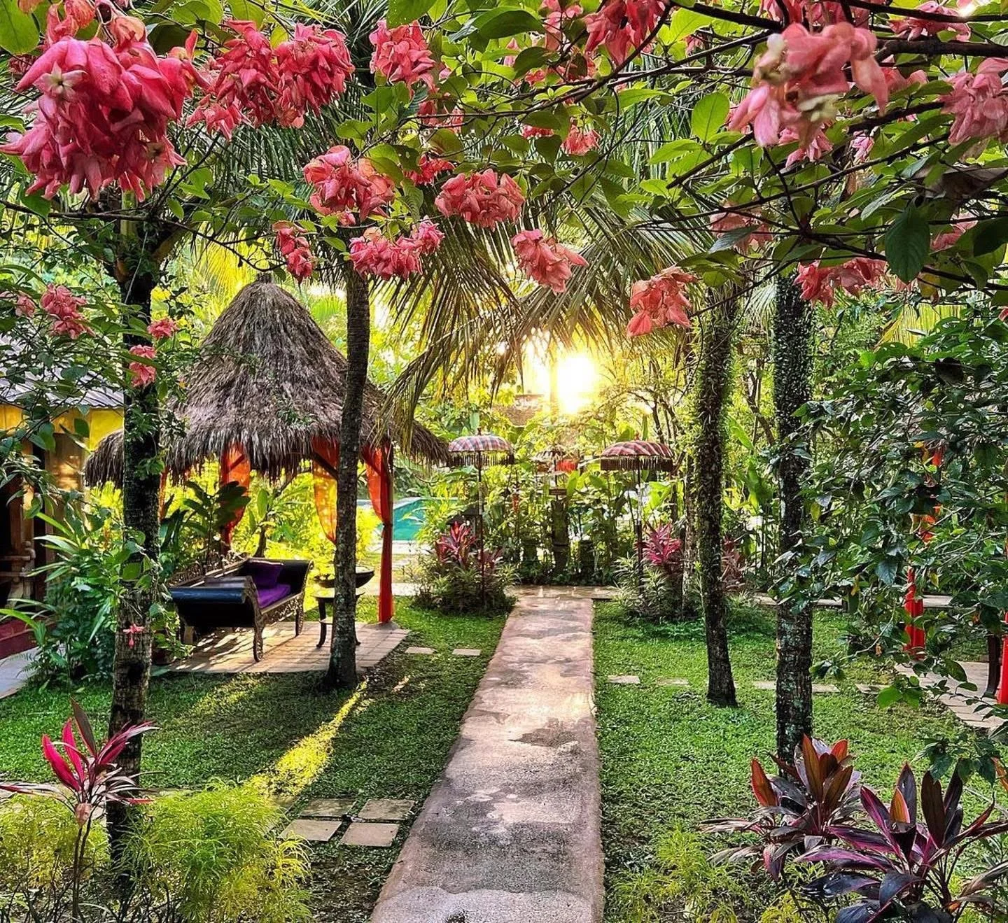 A lush tropical garden at sunset, featuring a stone pathway, colorful flowering plants, and small thatched huts with purple seating under pink umbrellas.