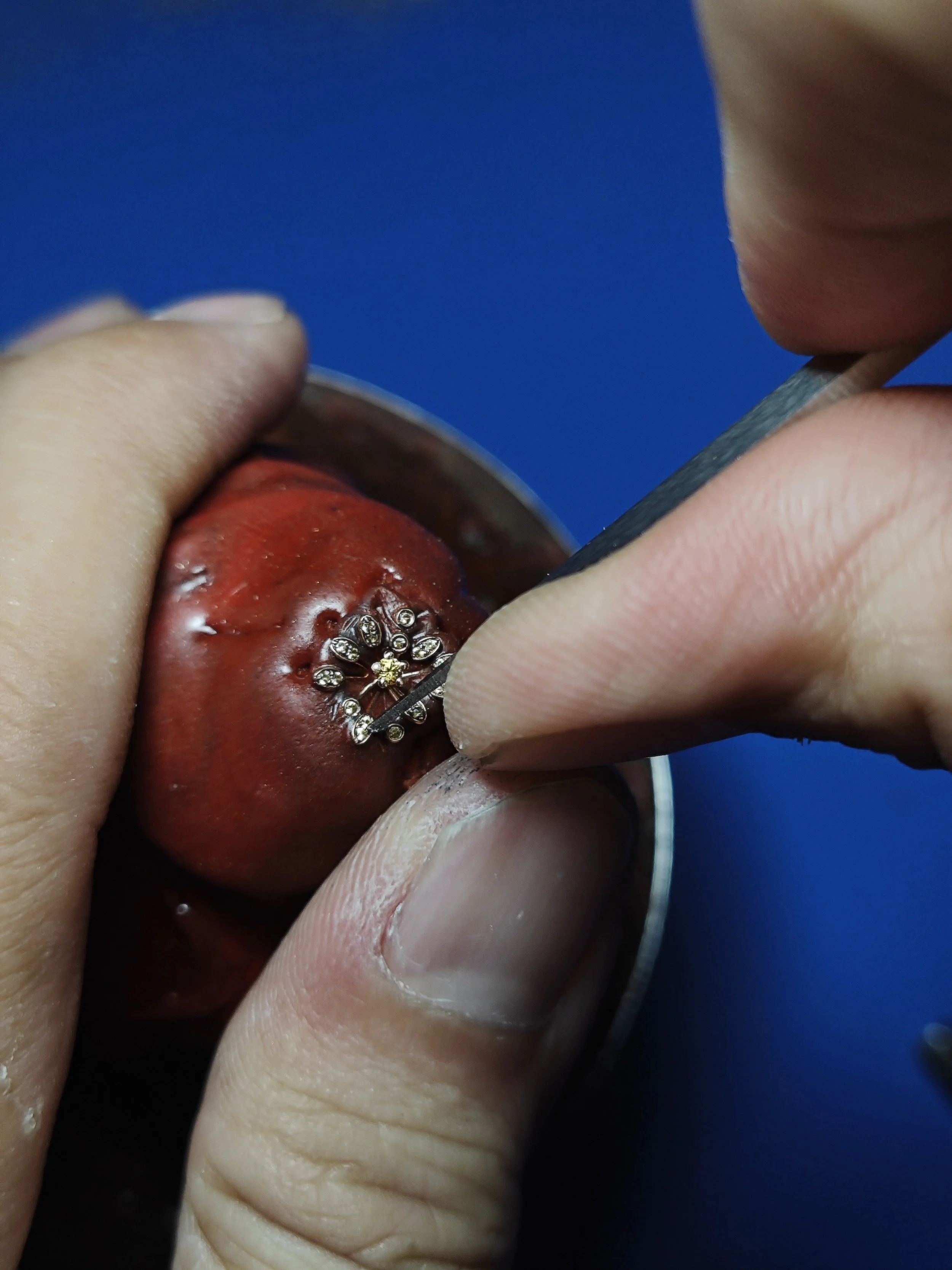 A close-up of hands holding a fruit with a tattoo. The person is inserting a needle with small rhinestones into the fruit, which appears to be a strawberry.