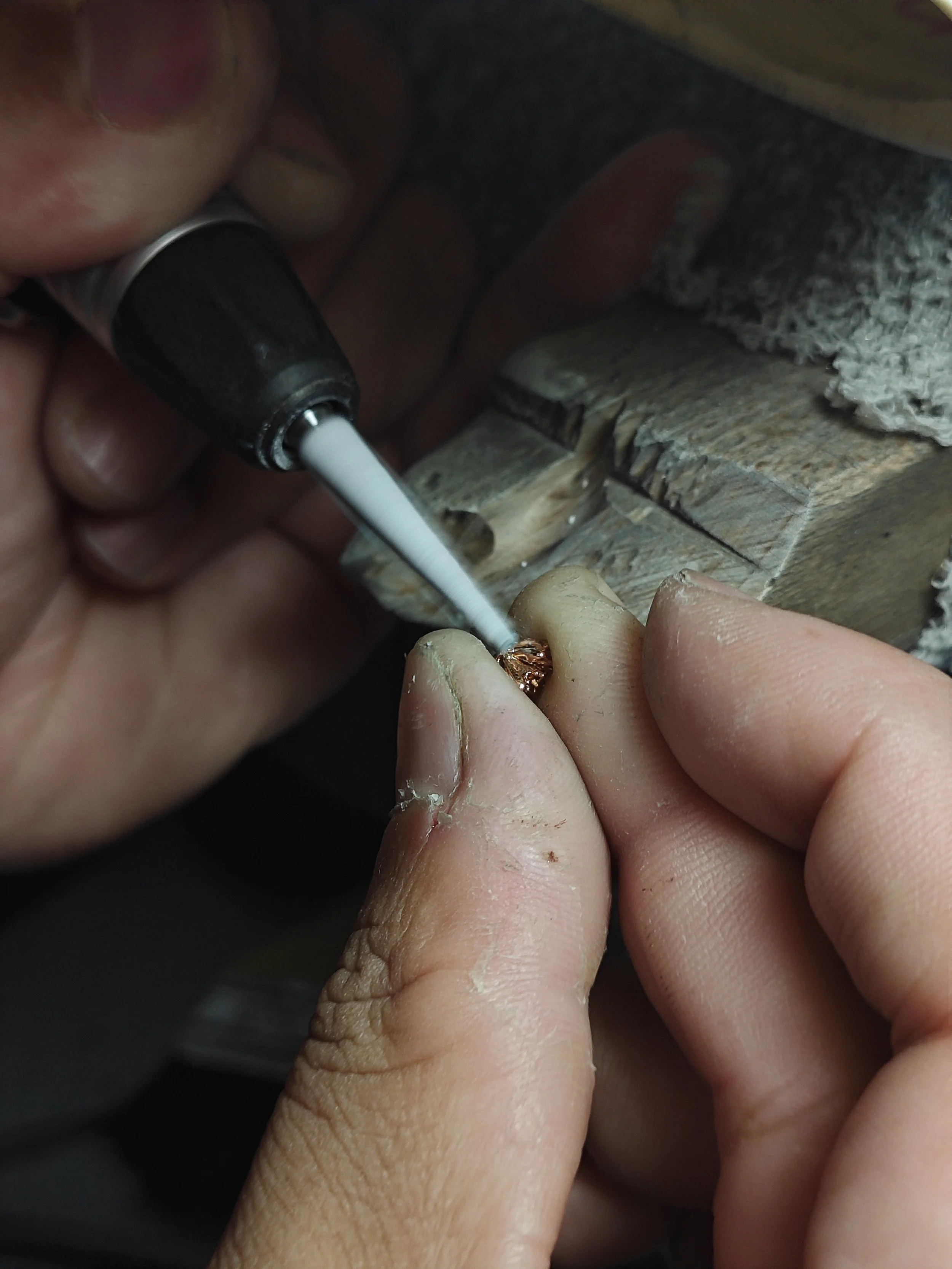 Close-up of hands working on a small, intricate project using a tool, with wood and rough material in the background.