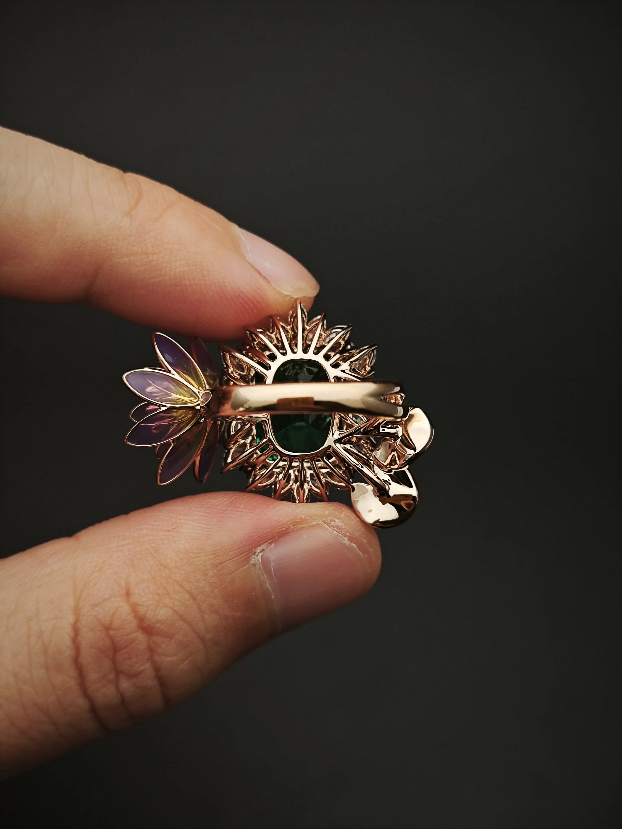 Close-up of a hand holding a gold ring with a sunflower-shaped design and a green gemstone at the center, against a dark background.