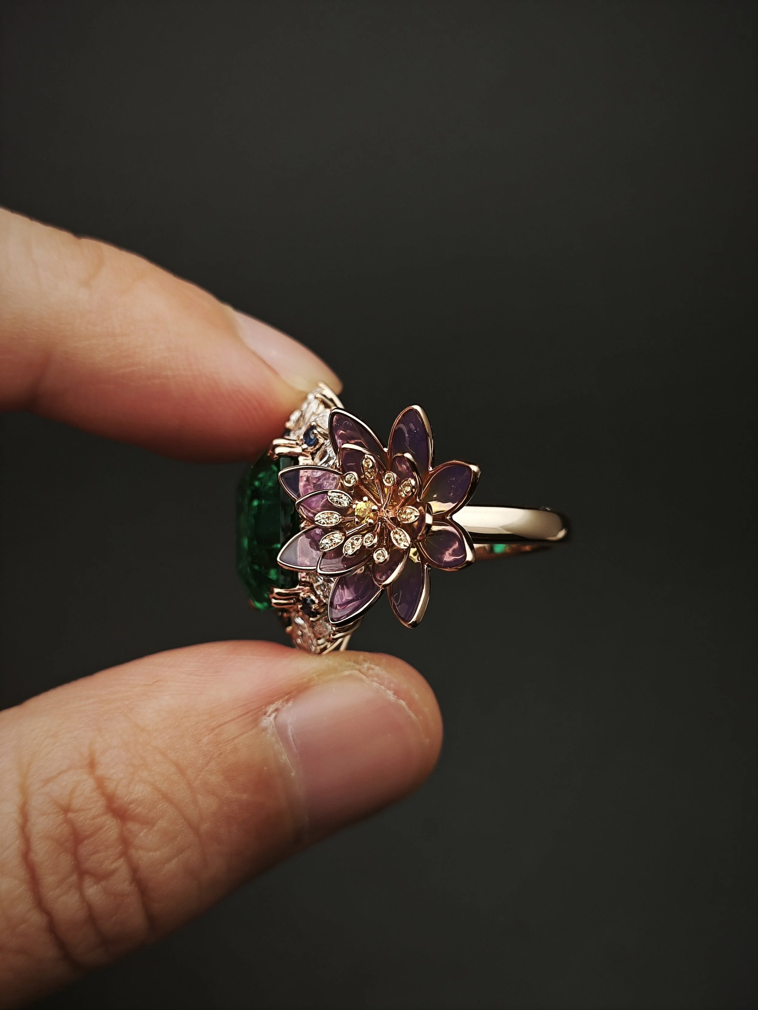 Close-up of a person's fingers holding a ring with a flower-shaped design featuring purple, pink, and white gemstones, set against a dark background.
