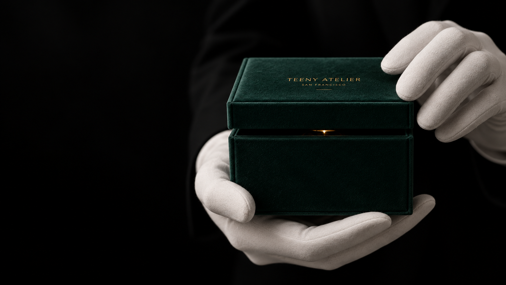 Person in white gloves holding a dark green jewelry box with gold lettering 'Teeny Atelier San Francisco' on the lid.