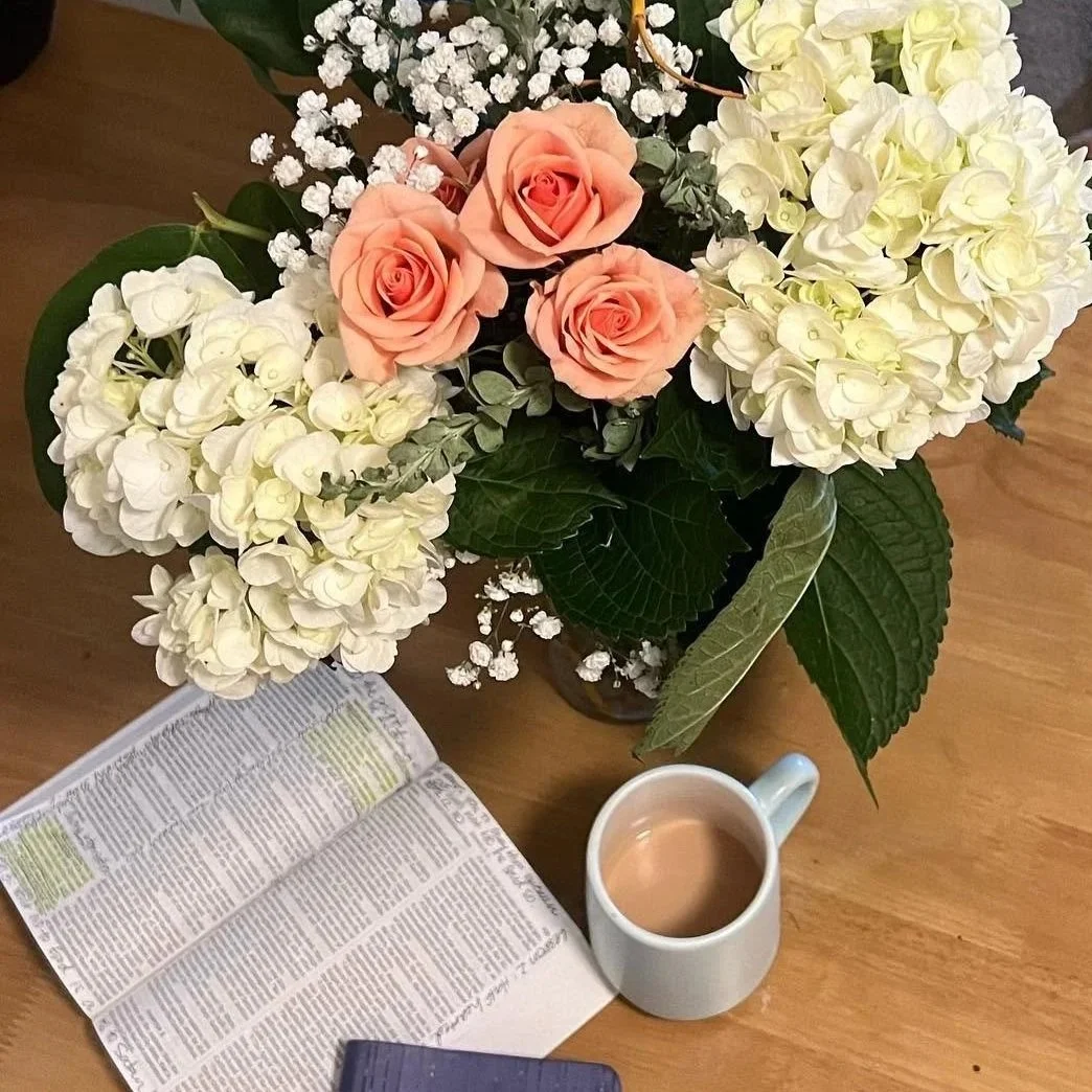 A bouquet of white hydrangeas, peach roses, and baby's breath on a wooden table. An open book, a white mug with coffee, and a small navy box are nearby.