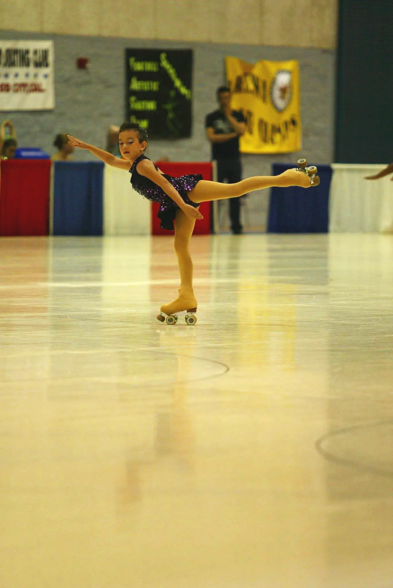 A young girl wearing a purple sparkly dress and beige roller skates performs a balance move on a skating rink. She is holding one arm out to the side and extending one leg behind her. In the background, a person, possibly a coach or judge, stands near a wall with banners and signs.