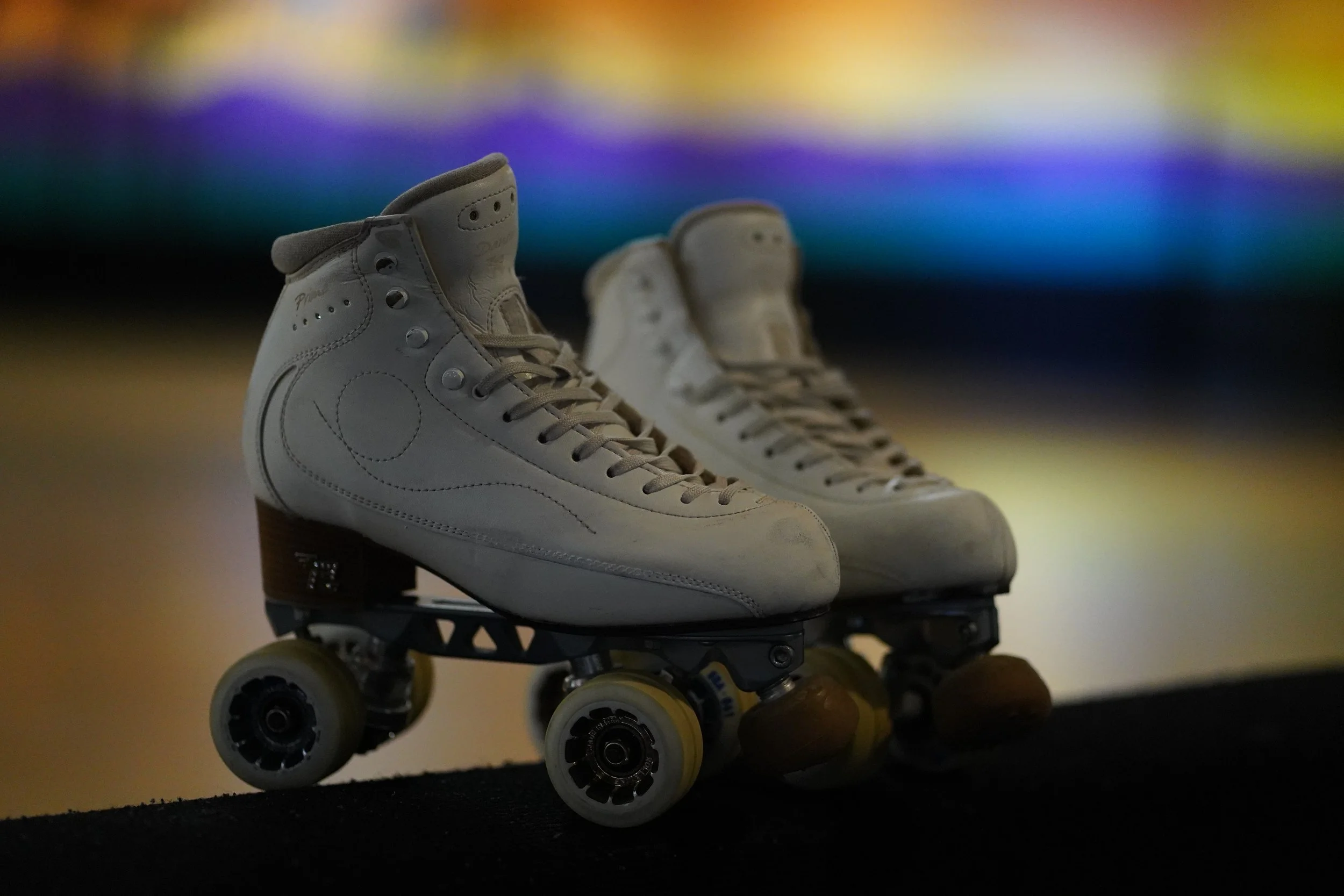 White vintage inline skate shoes with brown wheels and black trucks, placed on a black surface with a colorful blurry background.