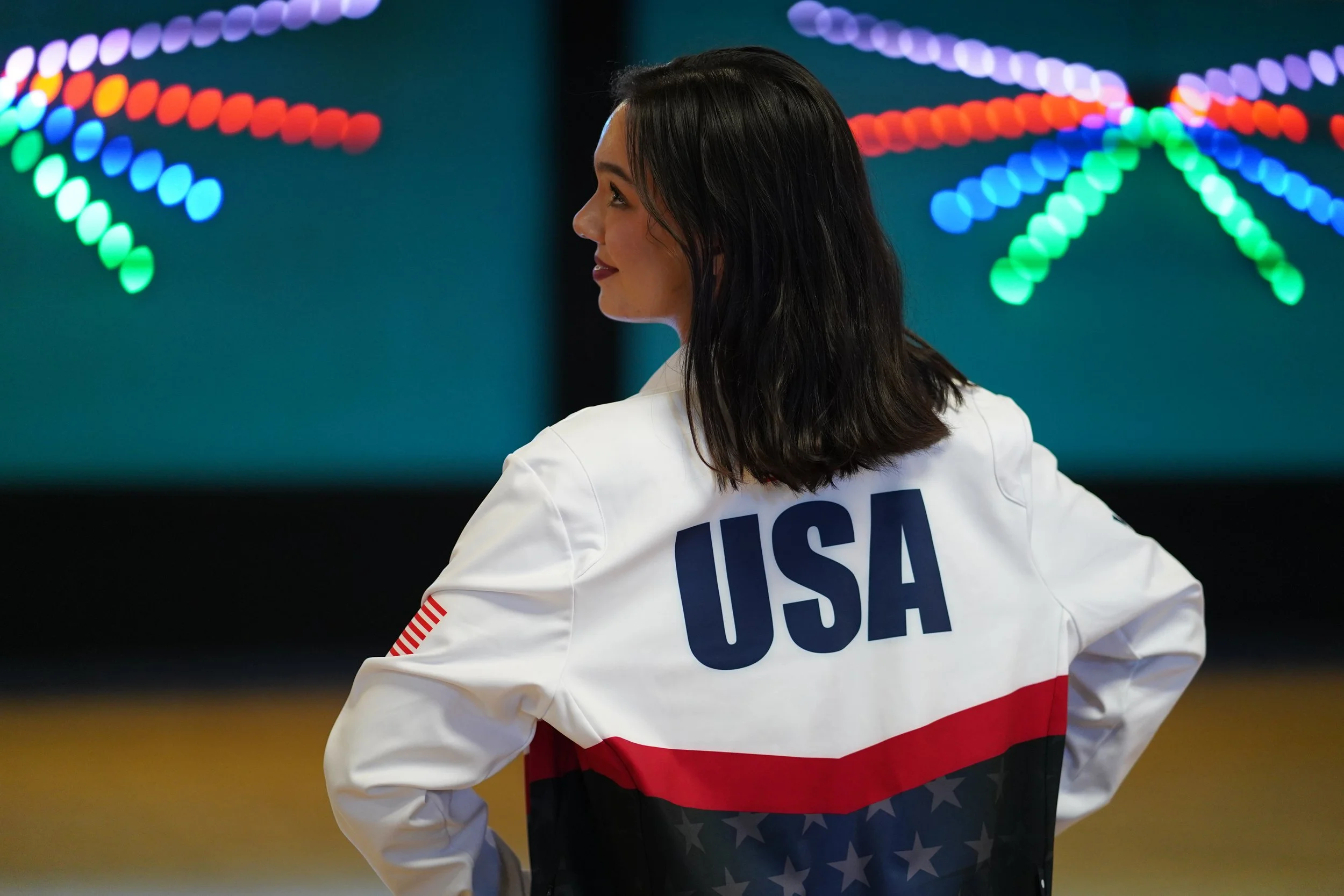 A woman wearing a jacket with 'USA' written on the back, standing in front of colorful neon lights arranged in a star pattern.