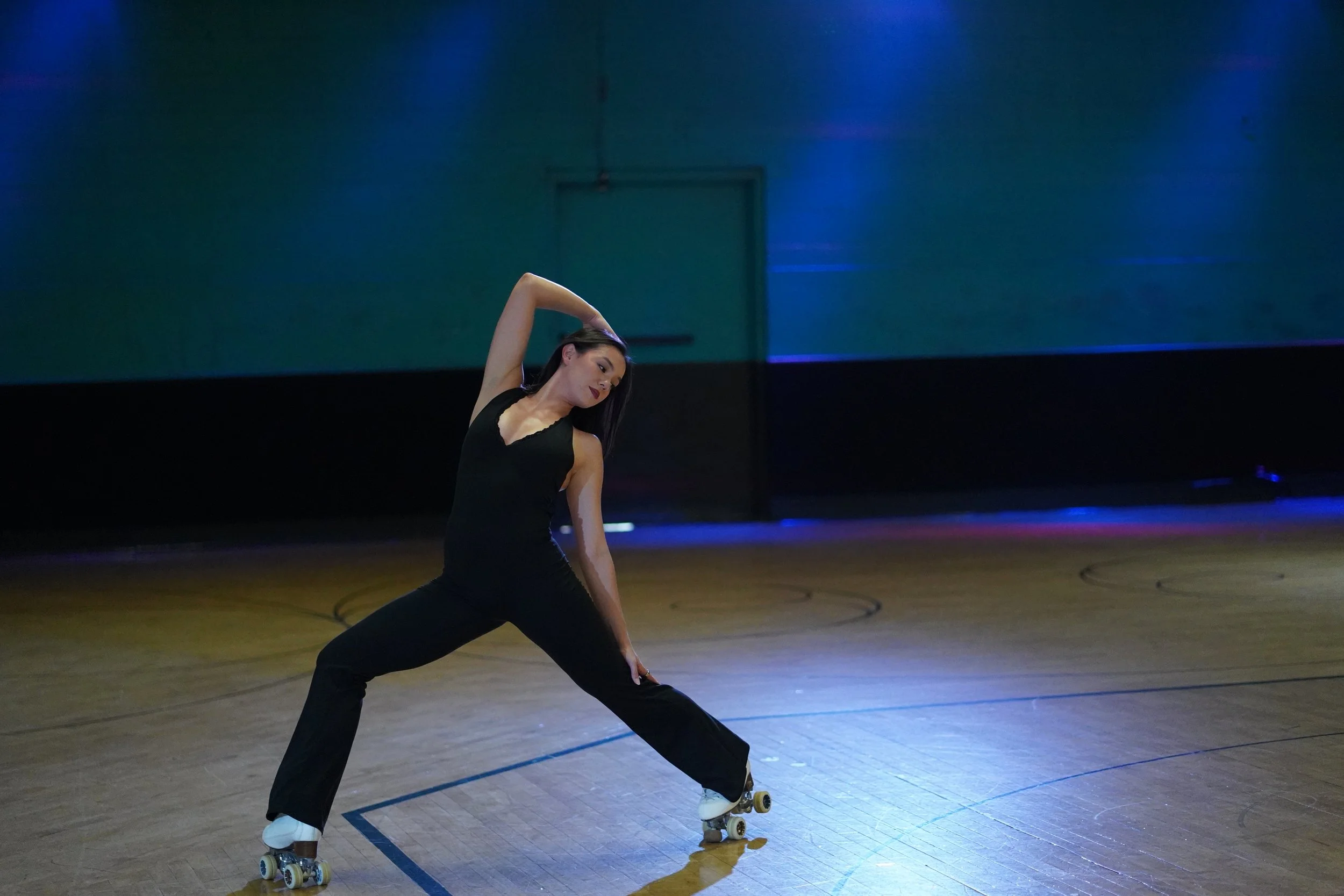 A woman in black clothing practicing roller skating dance moves in a dimly lit indoor dance studio.