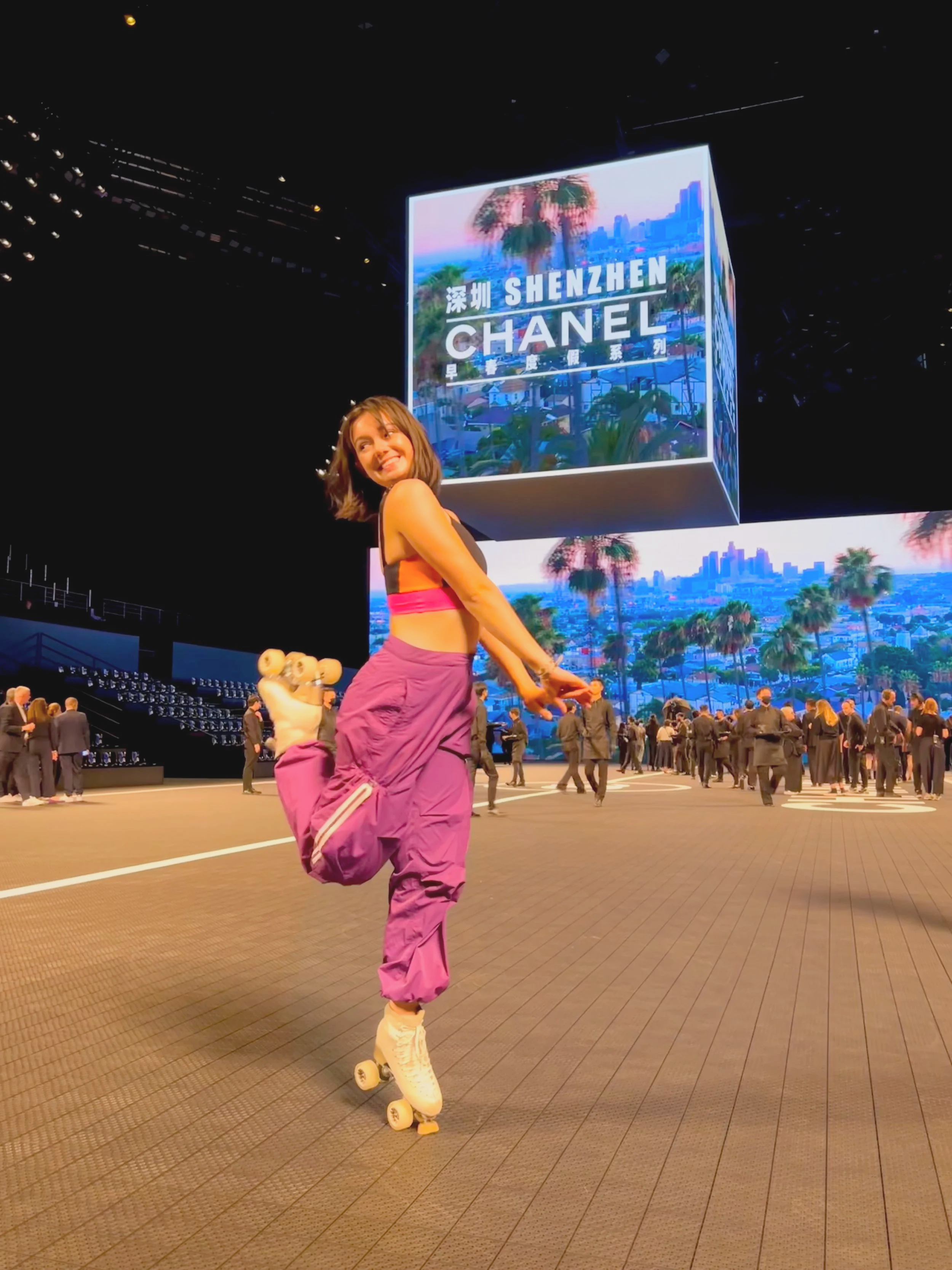A woman roller skating in front of a large digital billboard displaying images of Shenzhen, China, and the Chanel logo during an indoor event with many attendees.