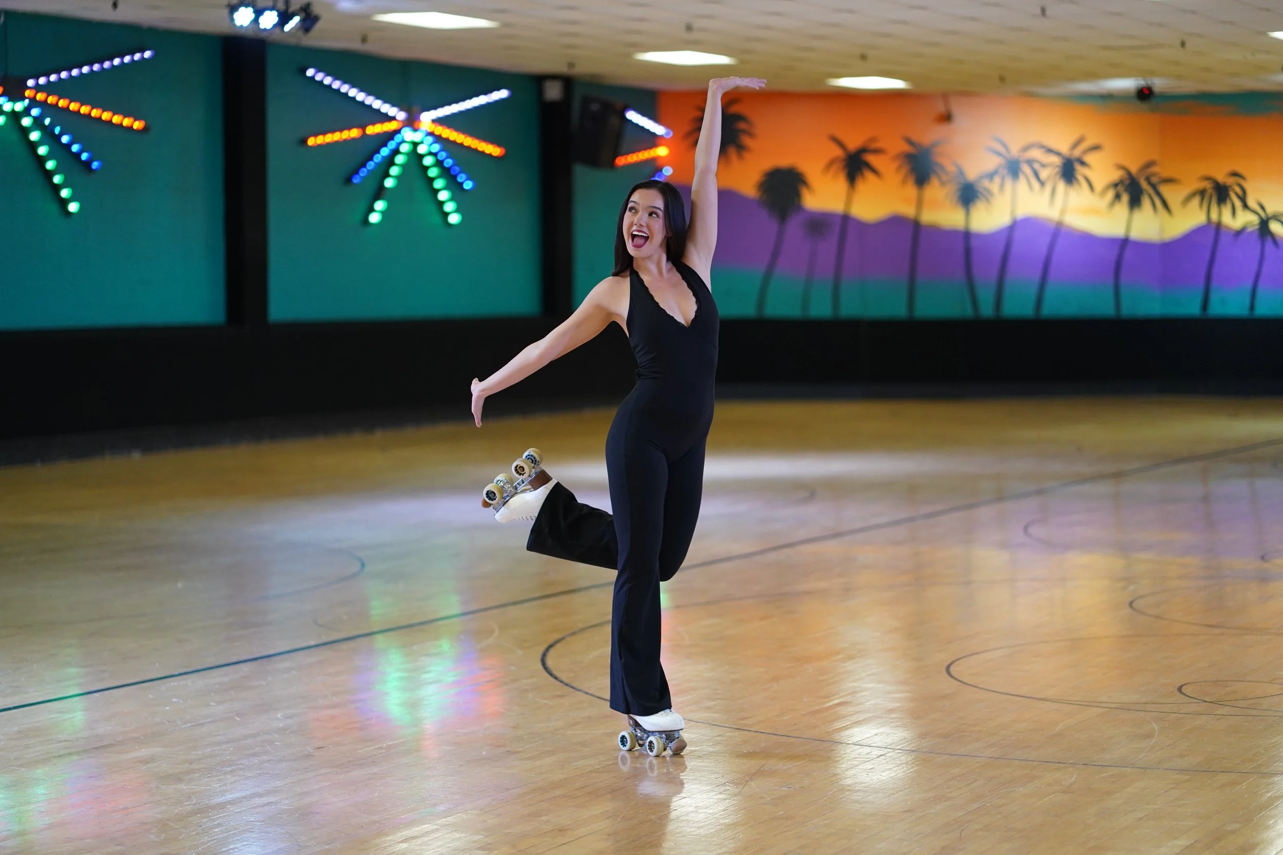A woman roller skating in a retro-themed roller rink with palm tree and sunset mural on the wall, decorated with colorful neon lights.