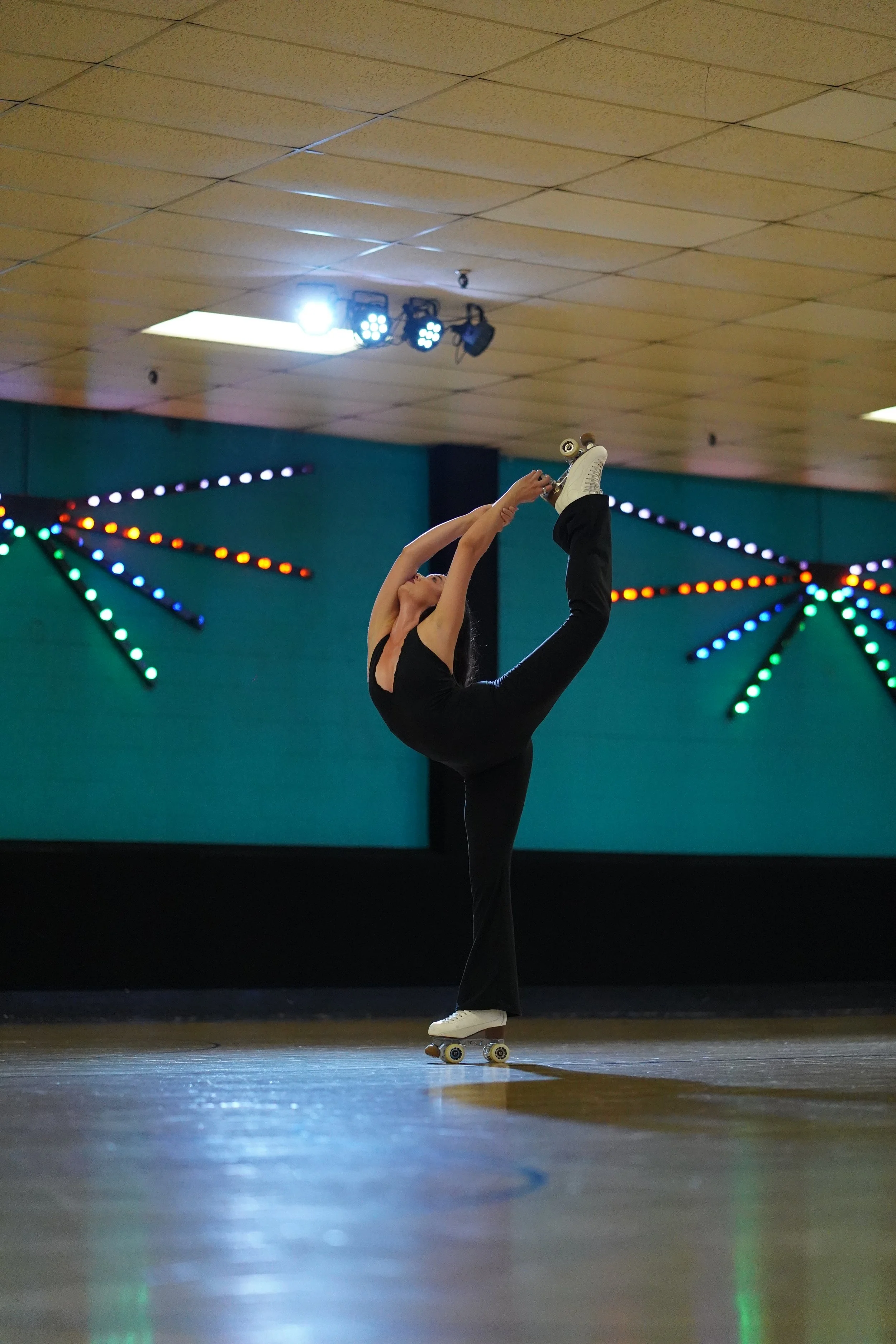 A person on roller skates in a black outfit performing an advanced backbend move on a wooden floor in an indoor roller skating rink, with brightly colored neon lights and a dark ceiling.