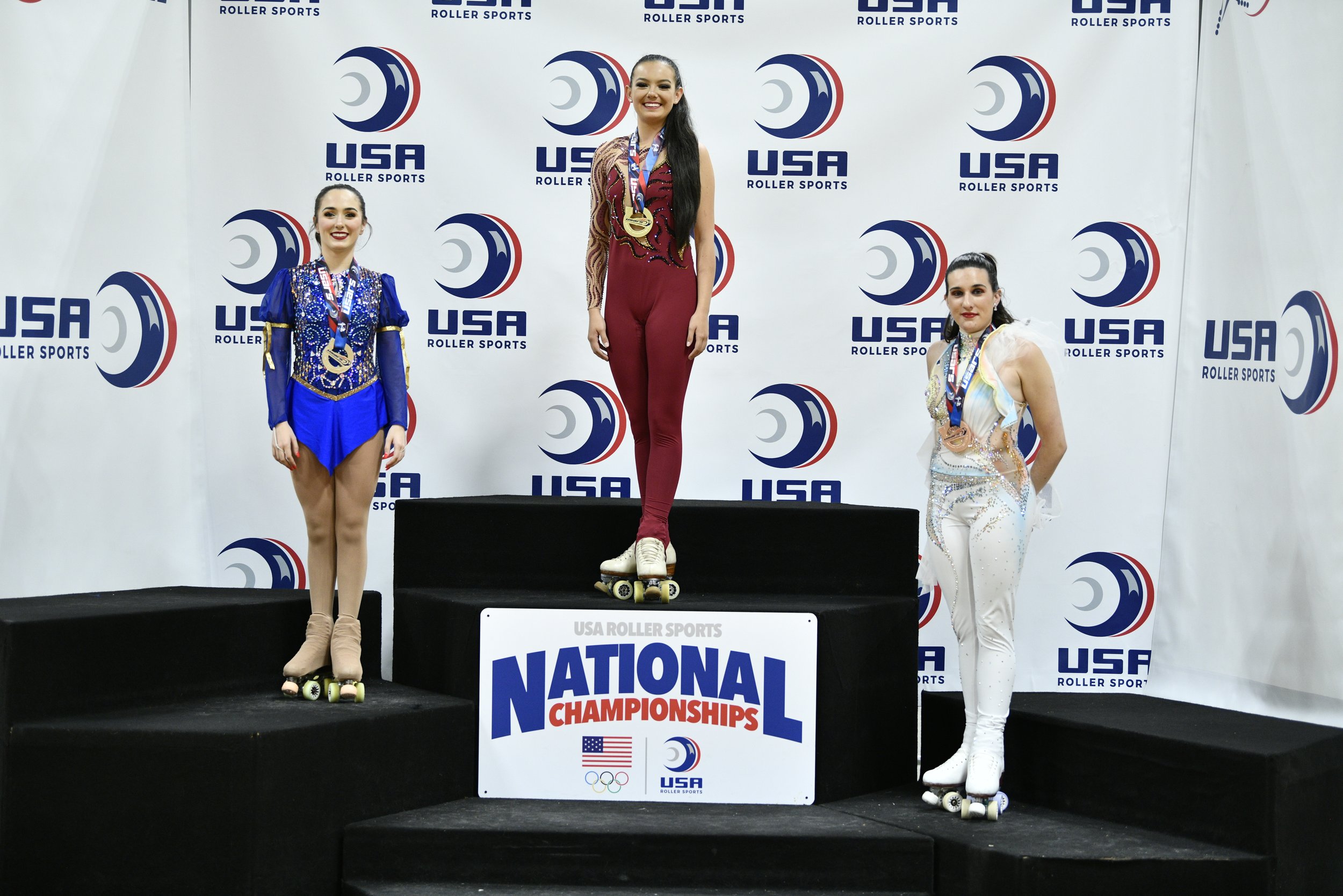 Three female roller skaters on a winners' podium at a USA Roller Sports National Championship, with medals around their necks, dressed in competition costumes, against a backdrop with USA Roller Sports logos. The skater in the center is on the top step, the other two are on either side.