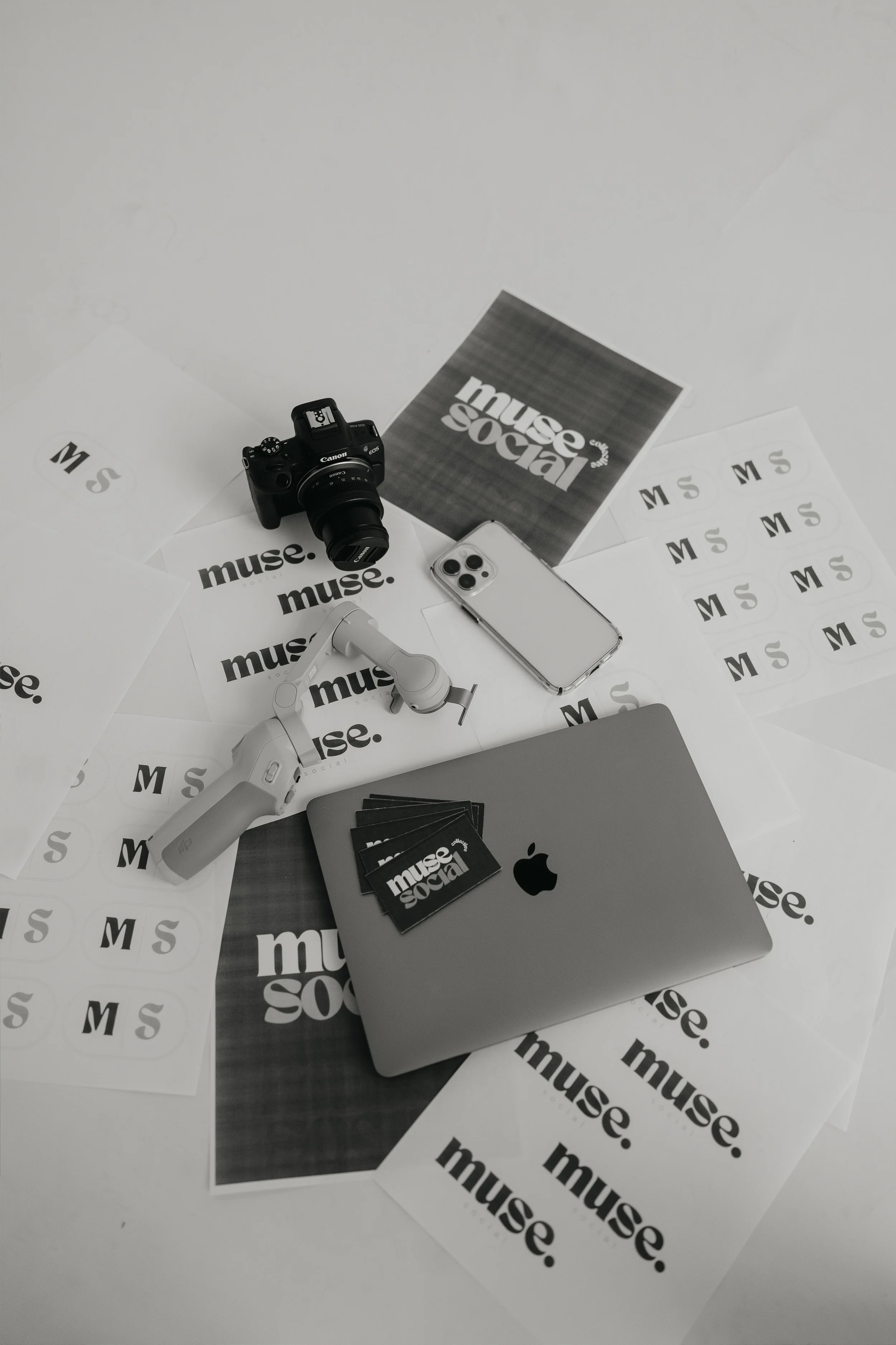 A flat lay of various items including a camera, smartphone, MacBook laptop, headphones, and printed stickers that say 'muse.social,' all arranged on a white surface covered with more stickers.