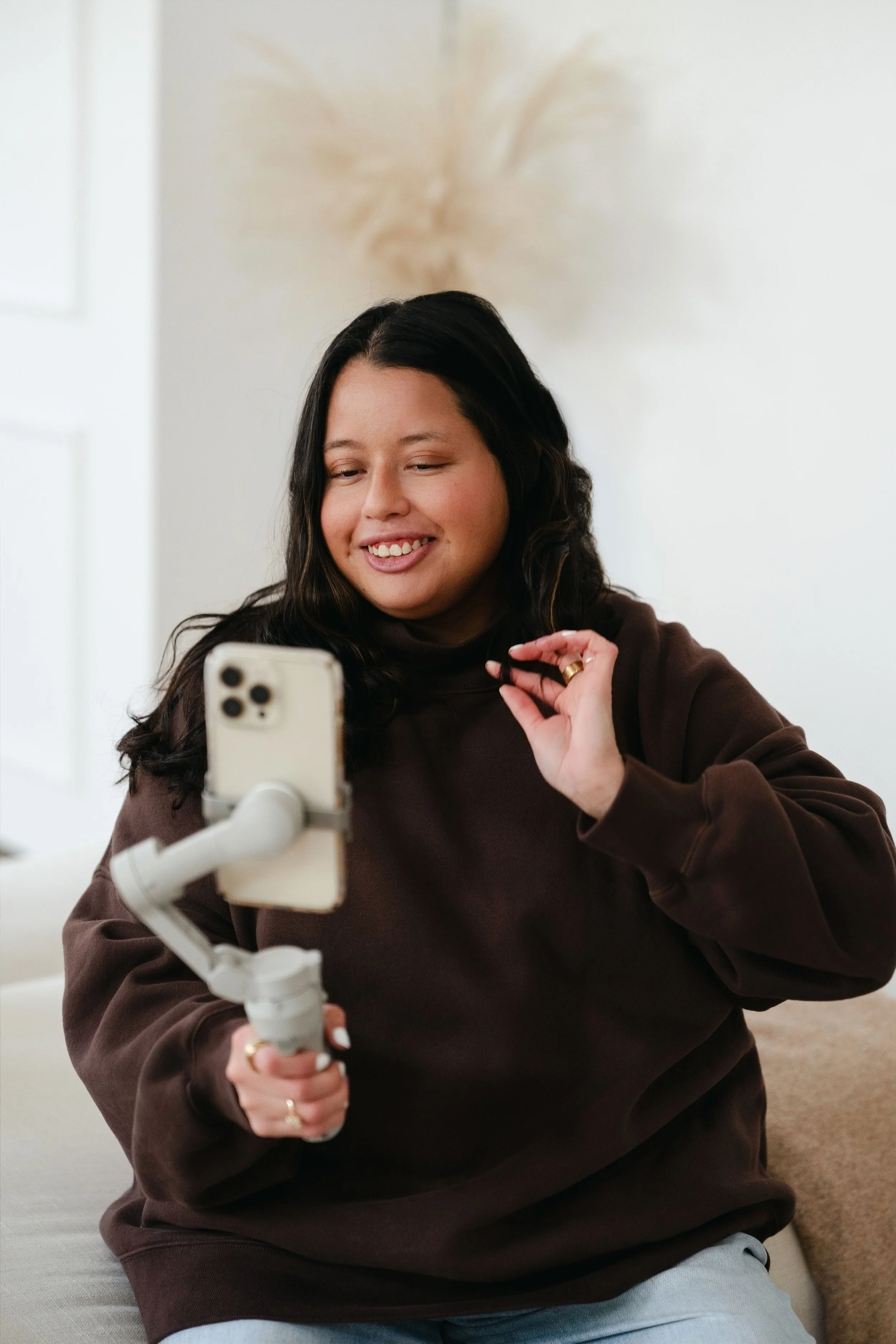 A woman with long dark hair smiling and taking a selfie with a smartphone mounted on a stabilizer. She is wearing a brown sweatshirt and sitting indoors with a neutral-colored background and a plant in the background.