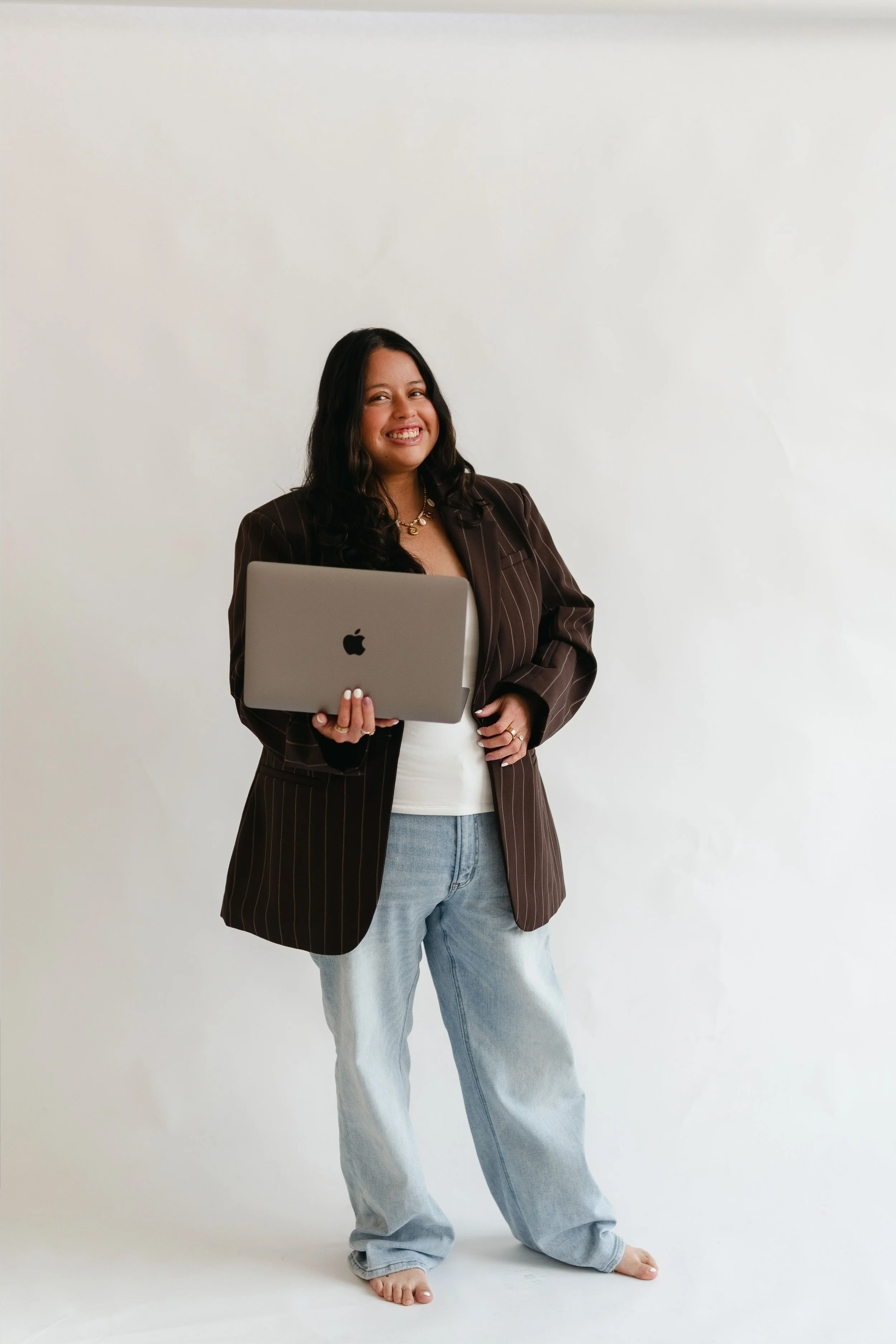 A woman with long dark hair smiling and holding a silver MacBook laptop, wearing a striped brown blazer, white top, and light blue jeans, standing barefoot against a plain white background.