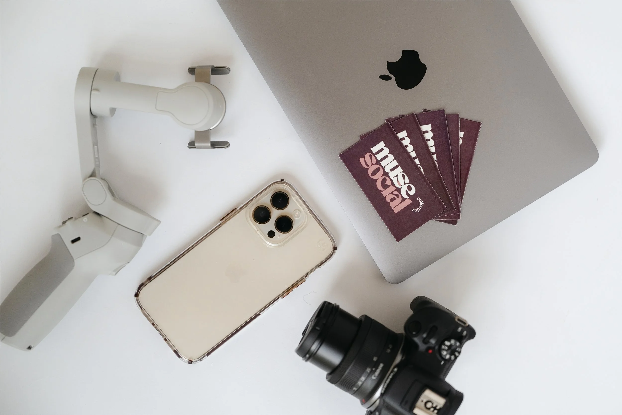 A flat lay of various electronic devices and accessories including a white mounted smartphone holder, an Apple MacBook with a black Apple logo, a bundle of five Marcus & Marcus gift cards, a white iPhone with triple-lens camera, a black DSLR camera, and a black camera lens, all on a white surface.