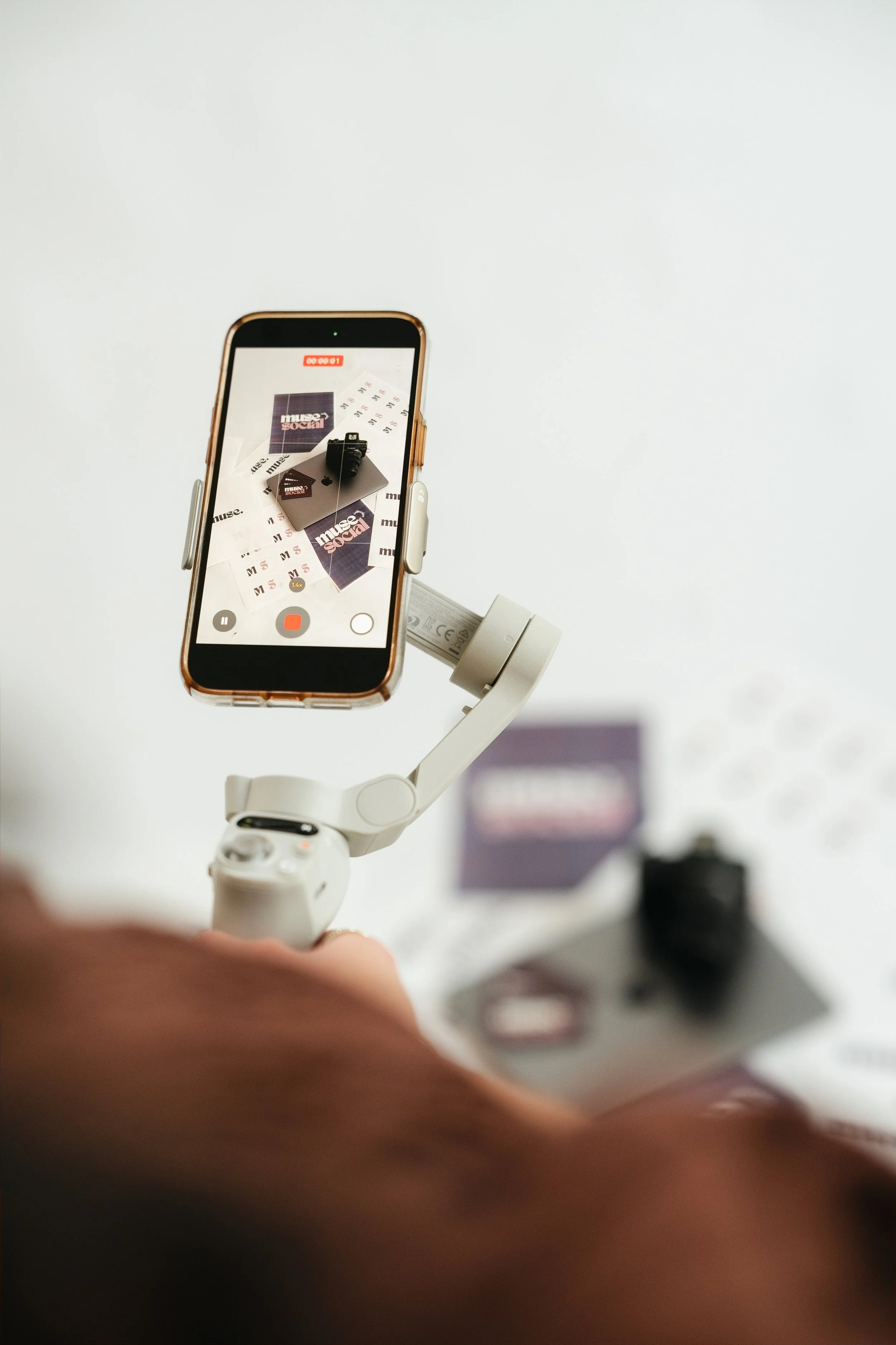 A smartphone mounted on a white gimbal stabilizer is capturing a top-down view of promotional materials that include stickers or cards with the words 'museum' and 'social' on a white surface.