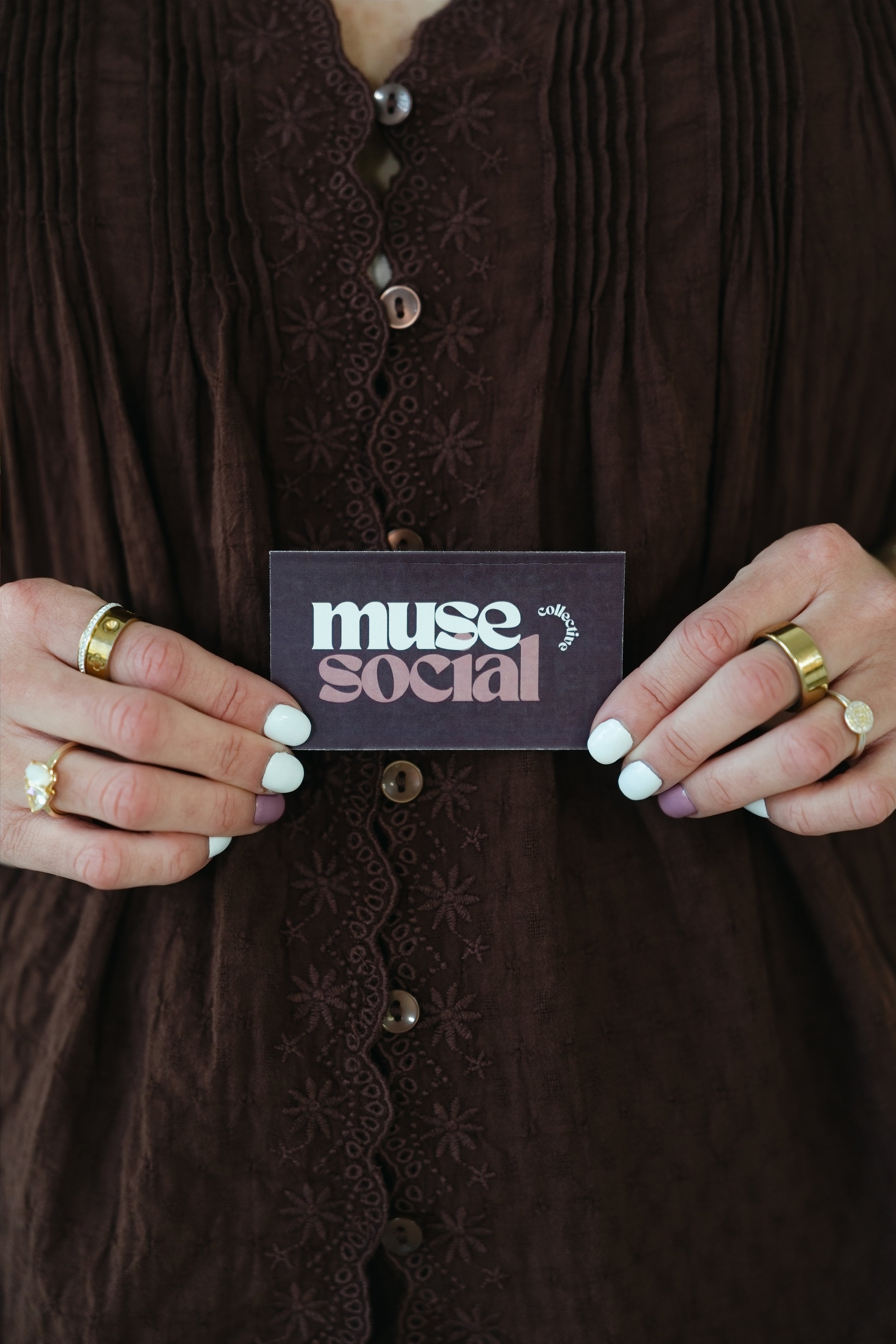 Person wearing a dark embroidered blouse holding a business card with the text 'muse social'.