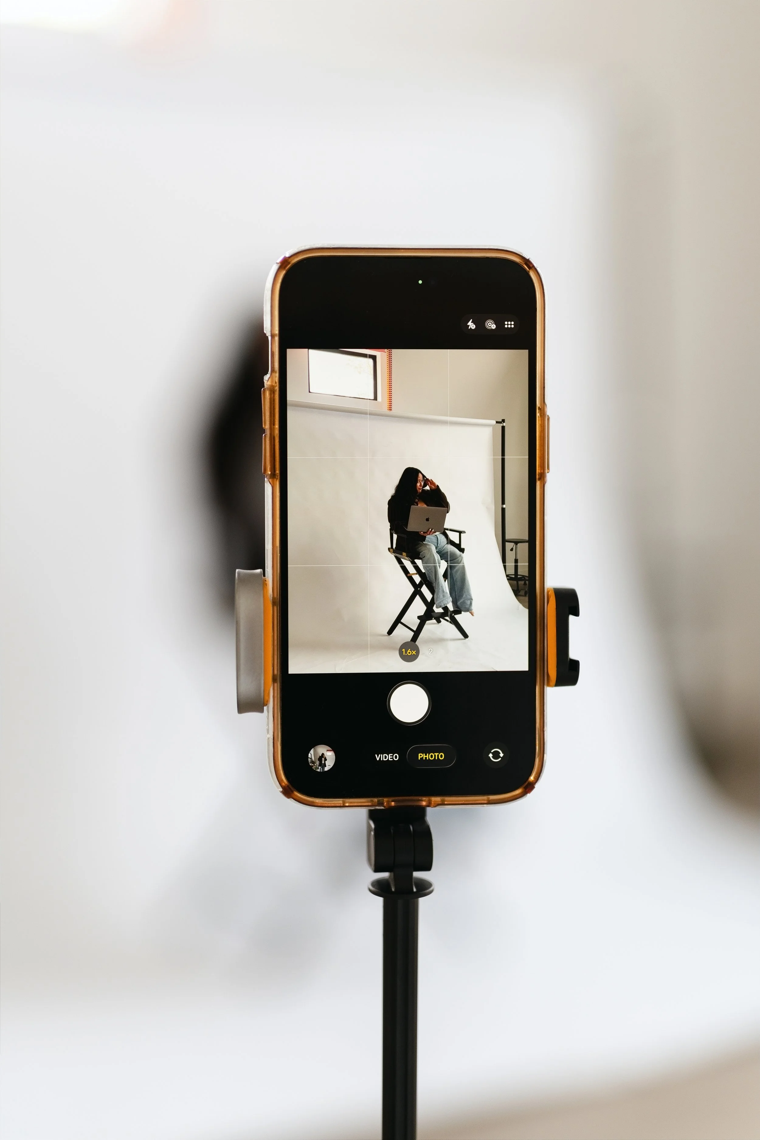 A smartphone mounted on a stand captures a woman sitting on a director's chair with a laptop, in a photography studio with a plain white background.