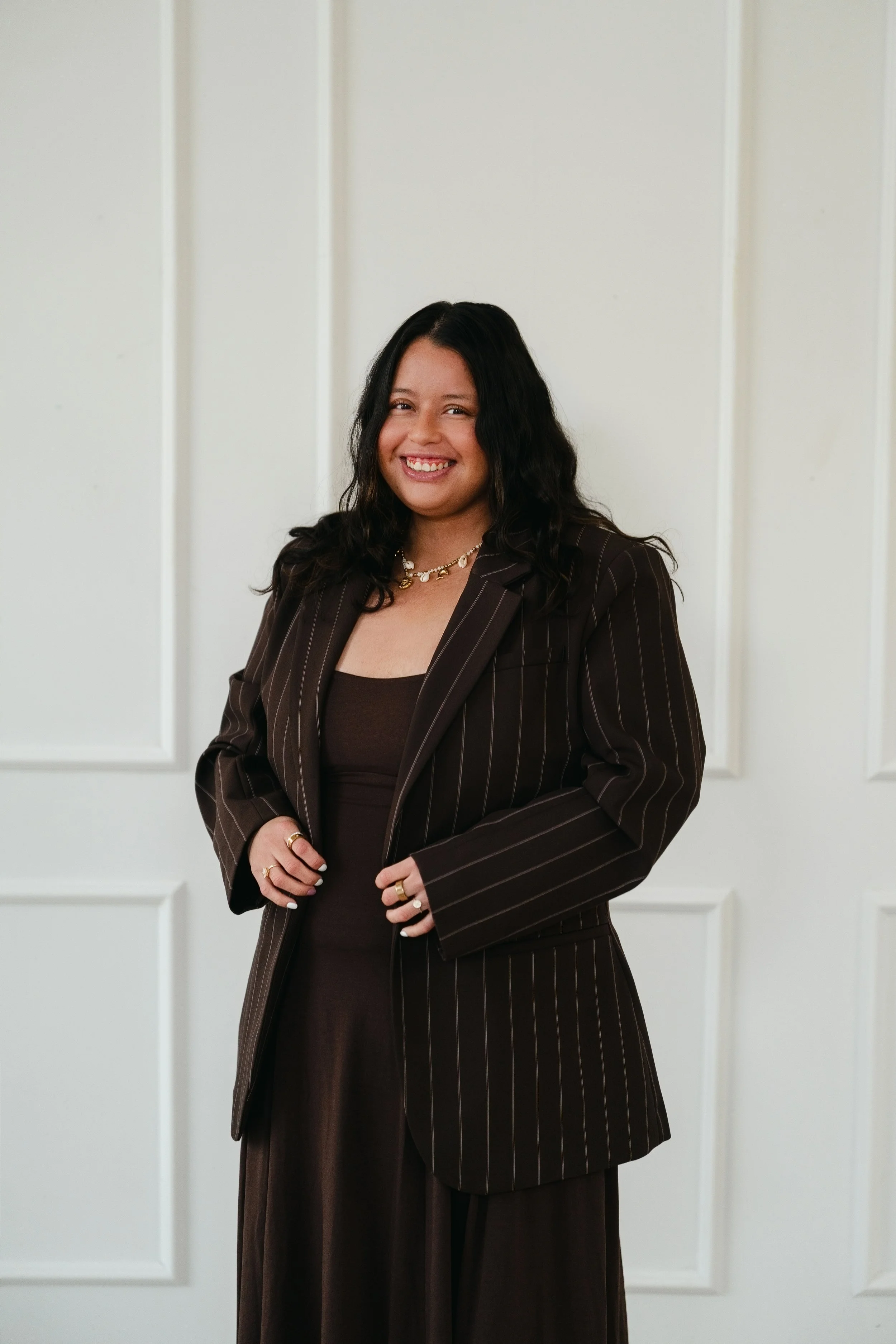A woman with black hair, wearing a brown pinstripe blazer and jewelry, smiling and posing against a white paneled wall.