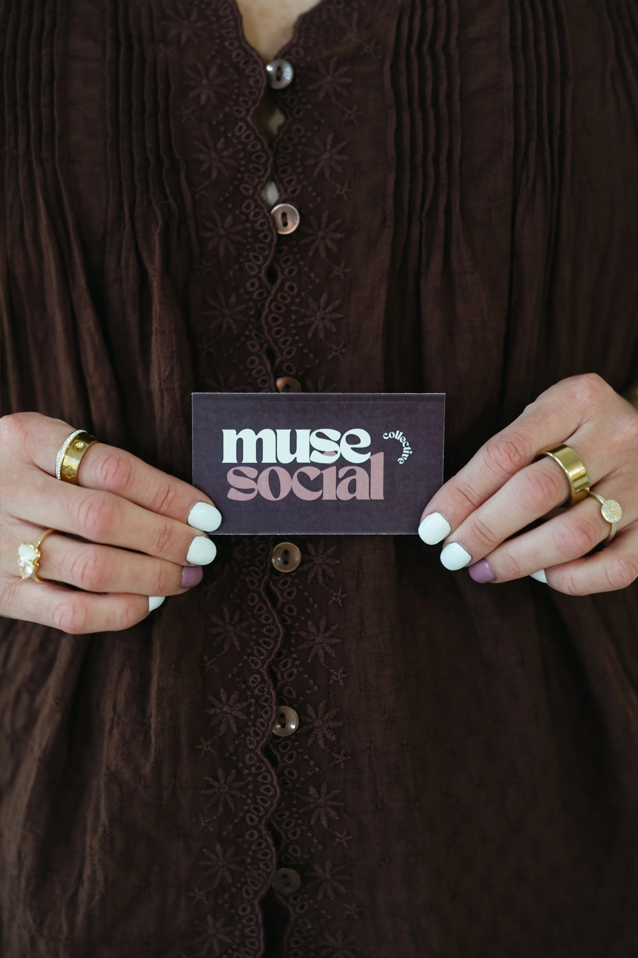 Person holding a business card with the text 'muse social' in front of their chest, wearing a dark embroidered blouse and rings on fingers.