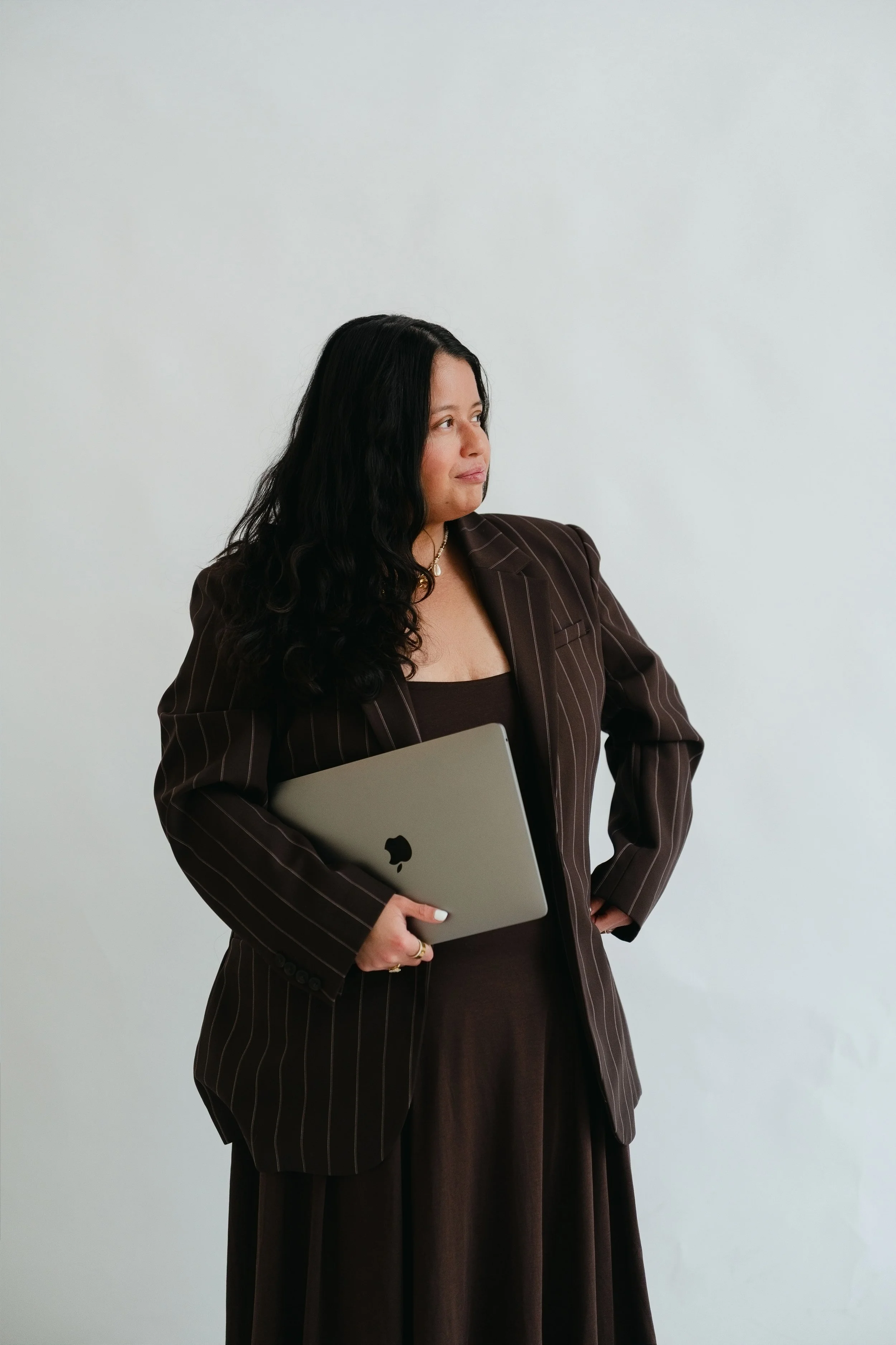 A woman with long black curly hair dressed in a brown pinstripe suit, holding a silver MacBook, standing against a plain white background, looking to her right.