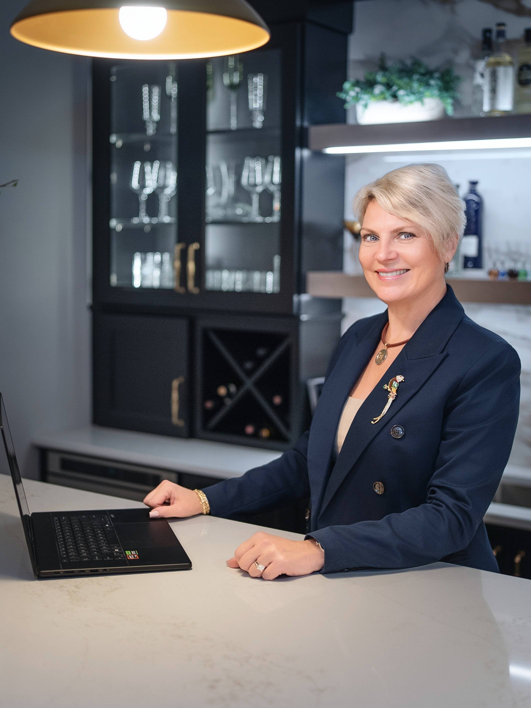 A woman with short blonde hair smiling while sitting at a table with a laptop in a modern kitchen or cafe setting, with shelves and glasses in the background.