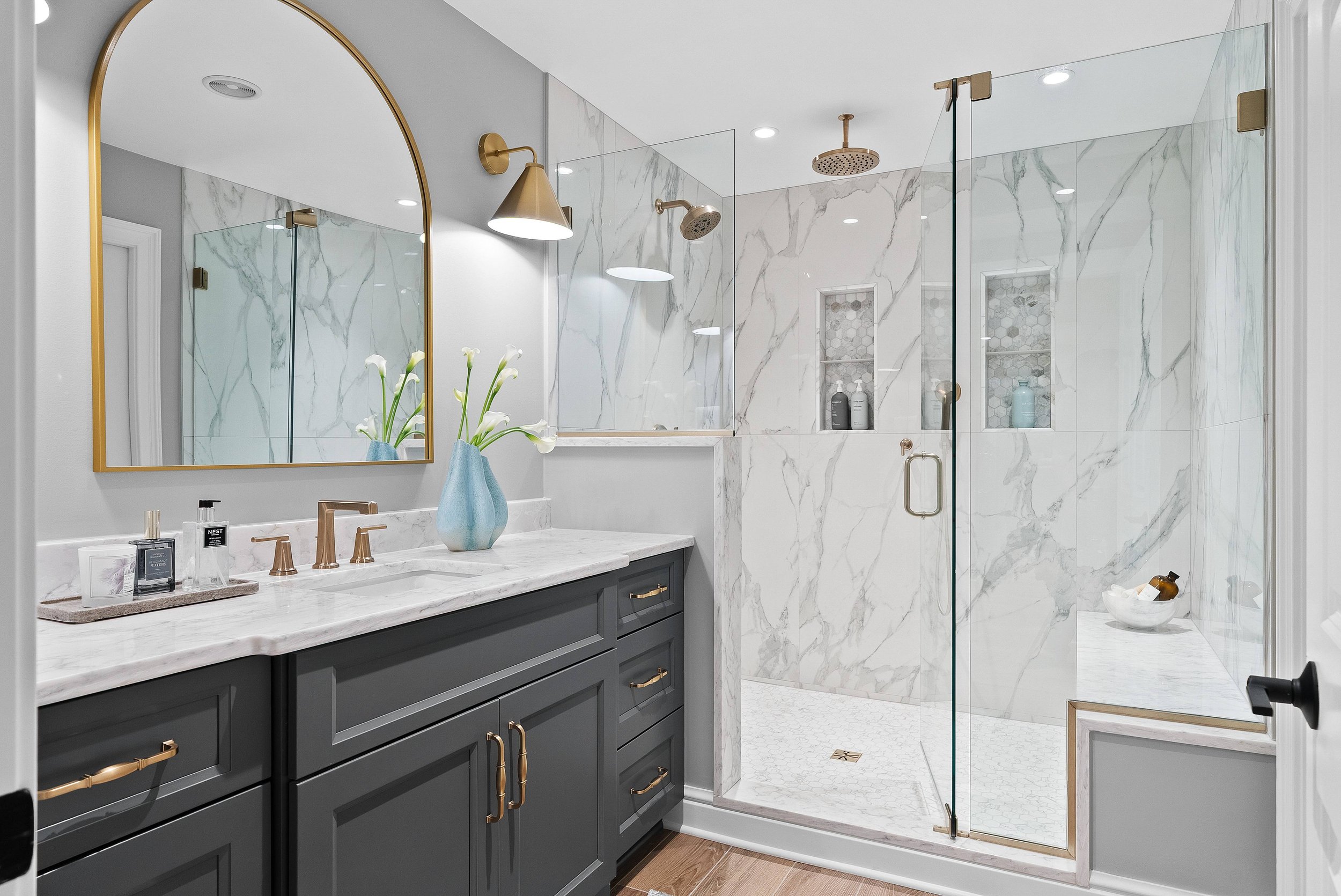 A modern bathroom with a white marble shower, gold fixtures, and a dark gray vanity with gold handles. A gold wall-mounted light, a large mirror with a gold frame, a vase with white calla lilies, and various toiletries are visible.
