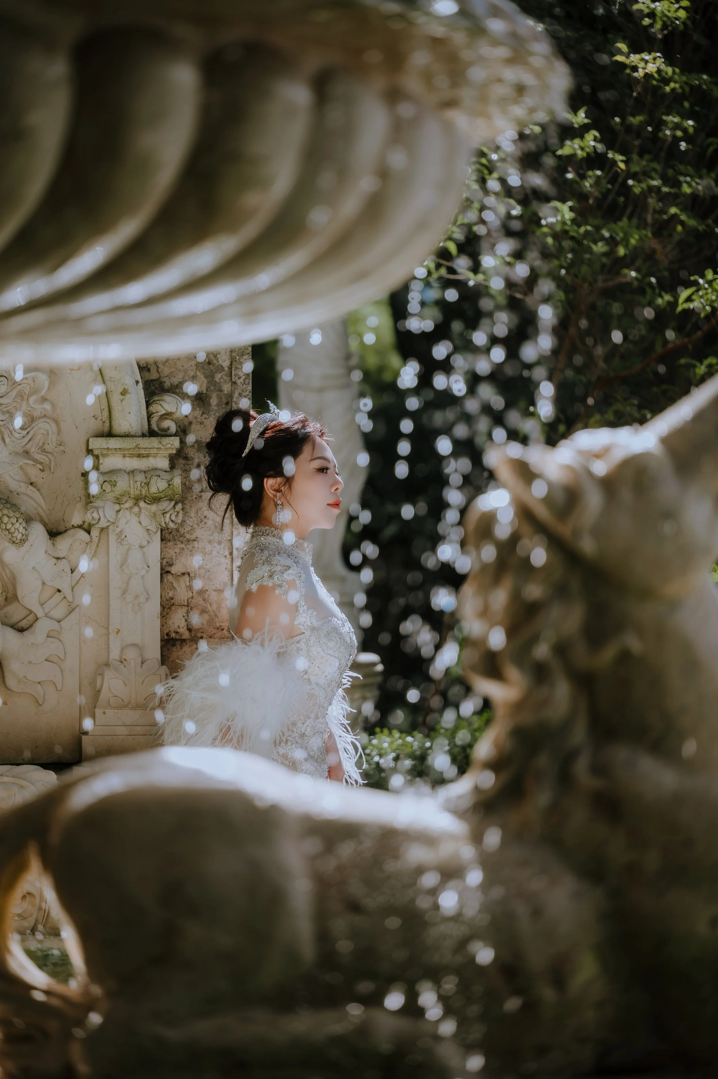 A bride in a white wedding gown with lace details and feather accents, wearing a tiara and dangling earrings, standing outdoors near a decorative stone wall, surrounded by greenery and dappled sunlight.