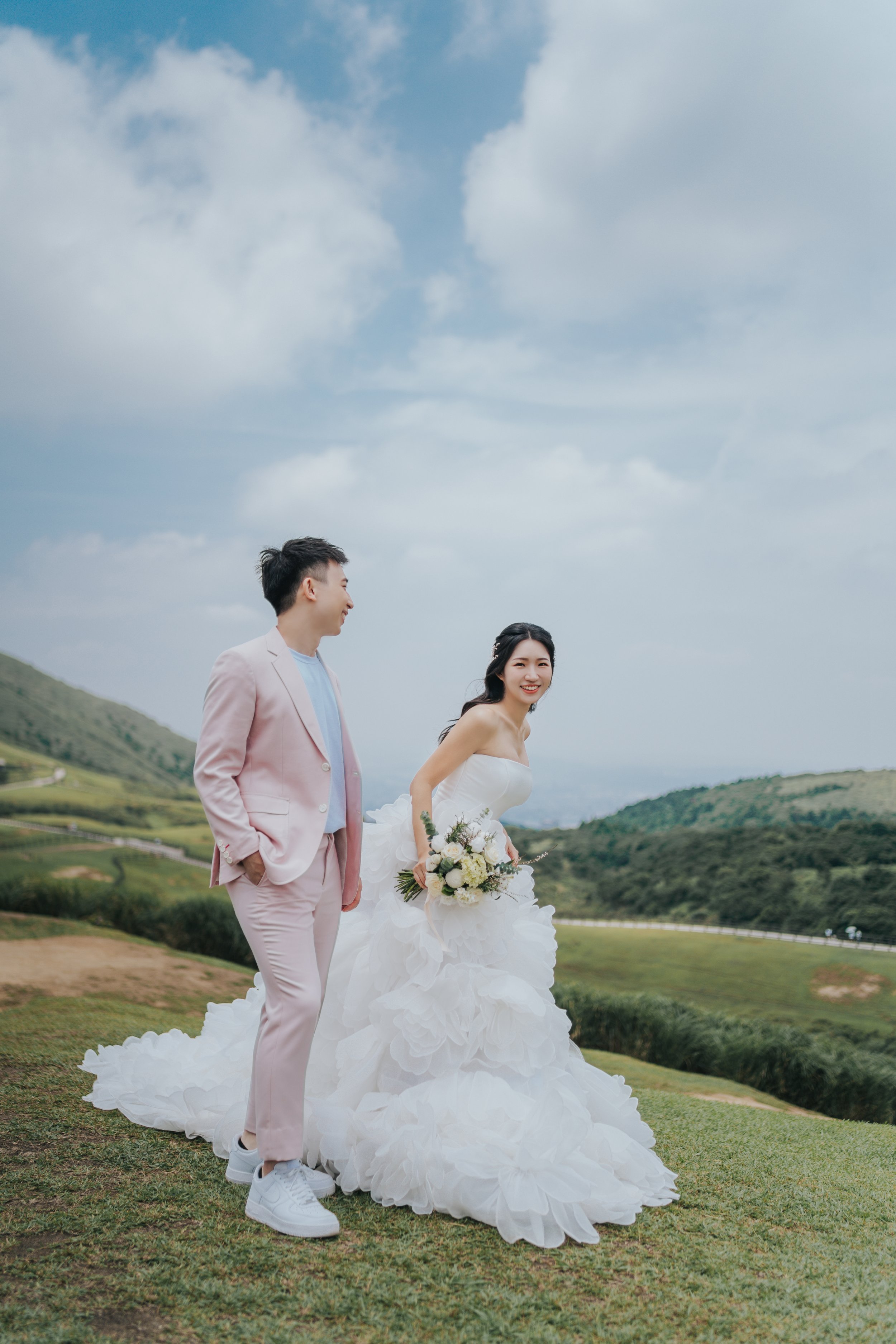 A bride and groom stand outdoors on a grassy hill, with a backdrop of rolling hills and a cloudy sky. The bride is wearing a strapless white wedding gown with a voluminous skirt and holding a bouquet, while the groom is dressed in a light pink suit with a light blue shirt and white sneakers. They are smiling and looking at each other.