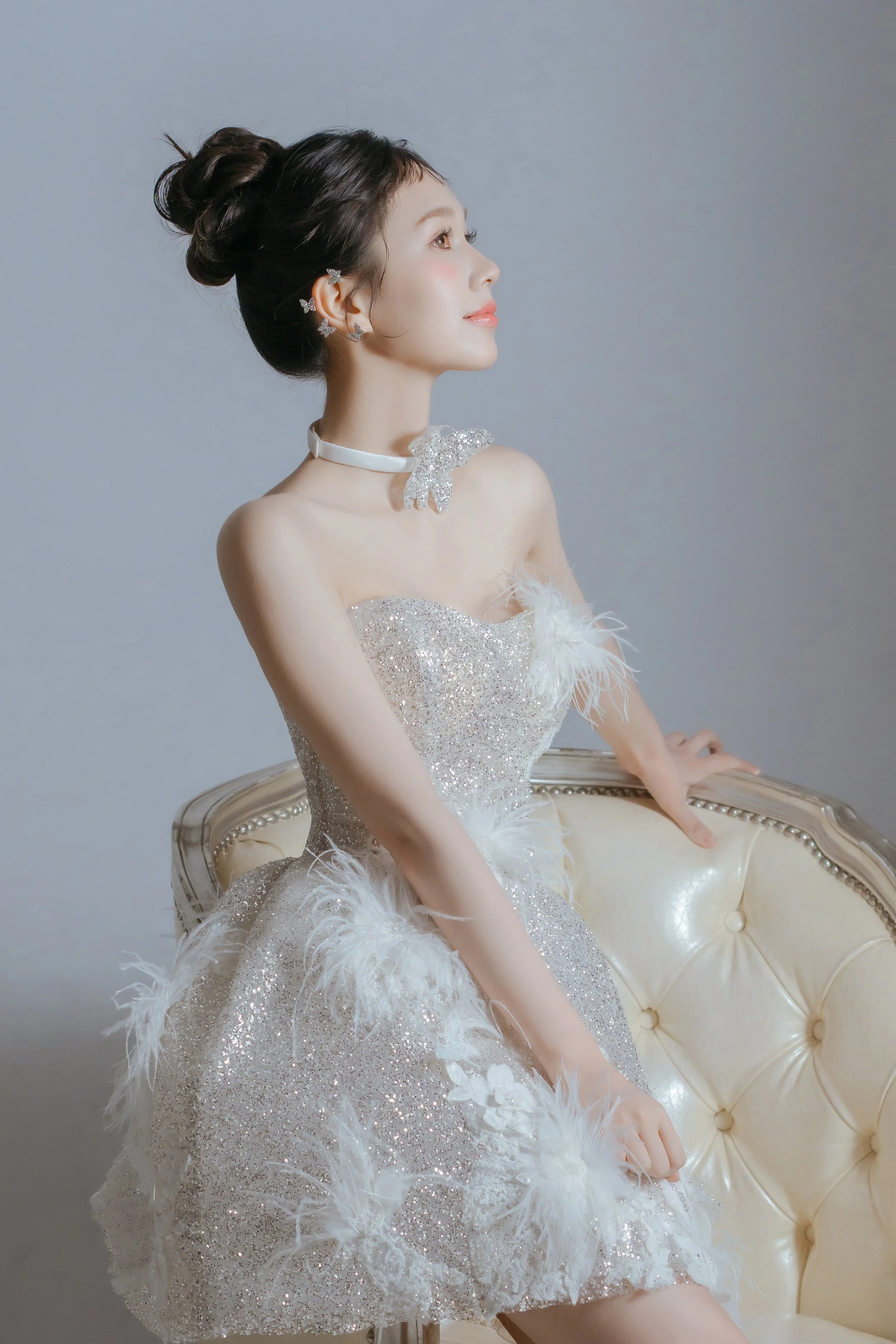 A woman with dark hair styled in an elegant updo, wearing butterfly-shaped earrings, a white choker with a bow, and a shimmering white dress with feathers, posed sitting on a vintage chair with a tufted leather backrest.