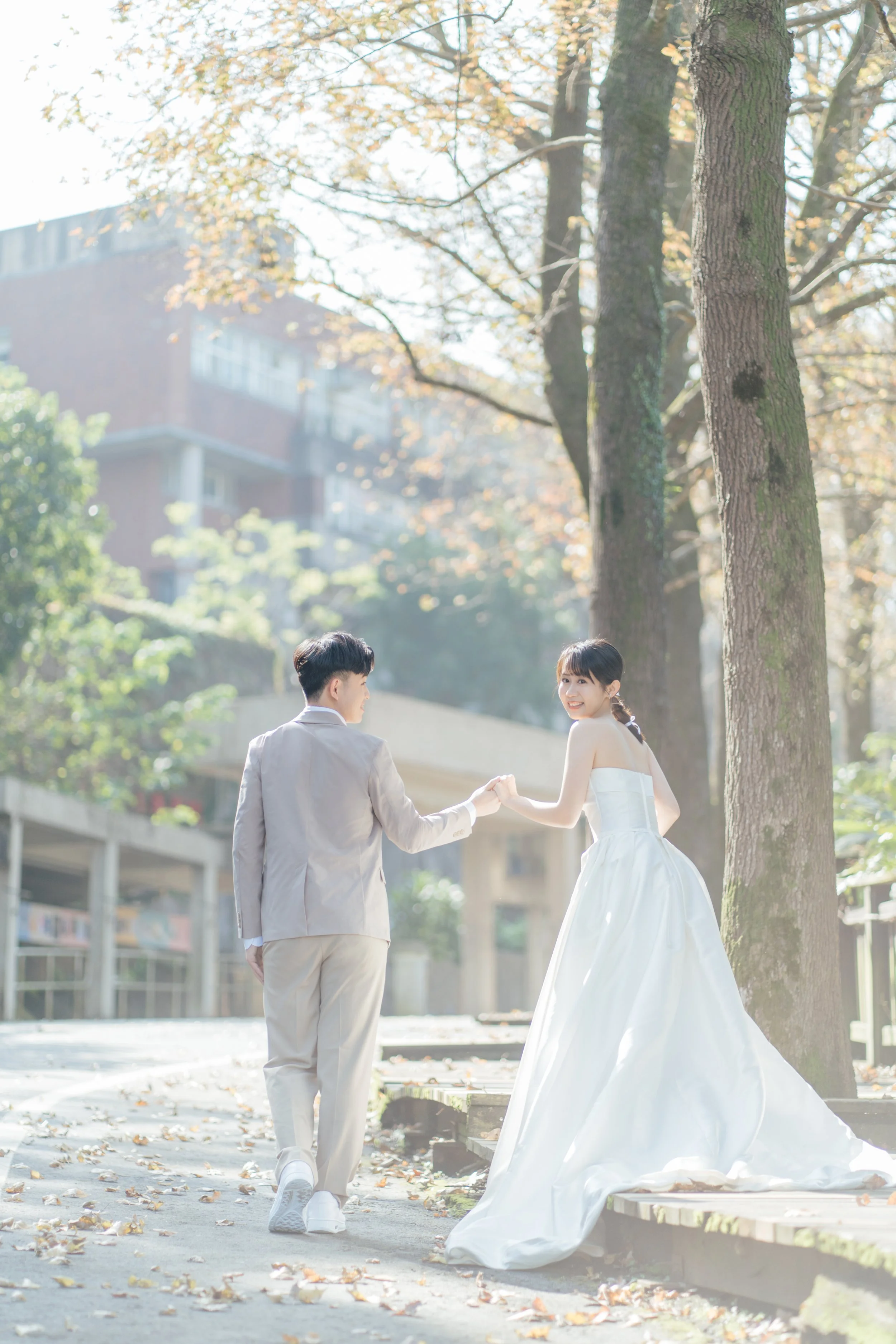 A couple dressed in wedding attire holding hands outdoors in a park, with trees and a building in the background on a sunny autumn day.