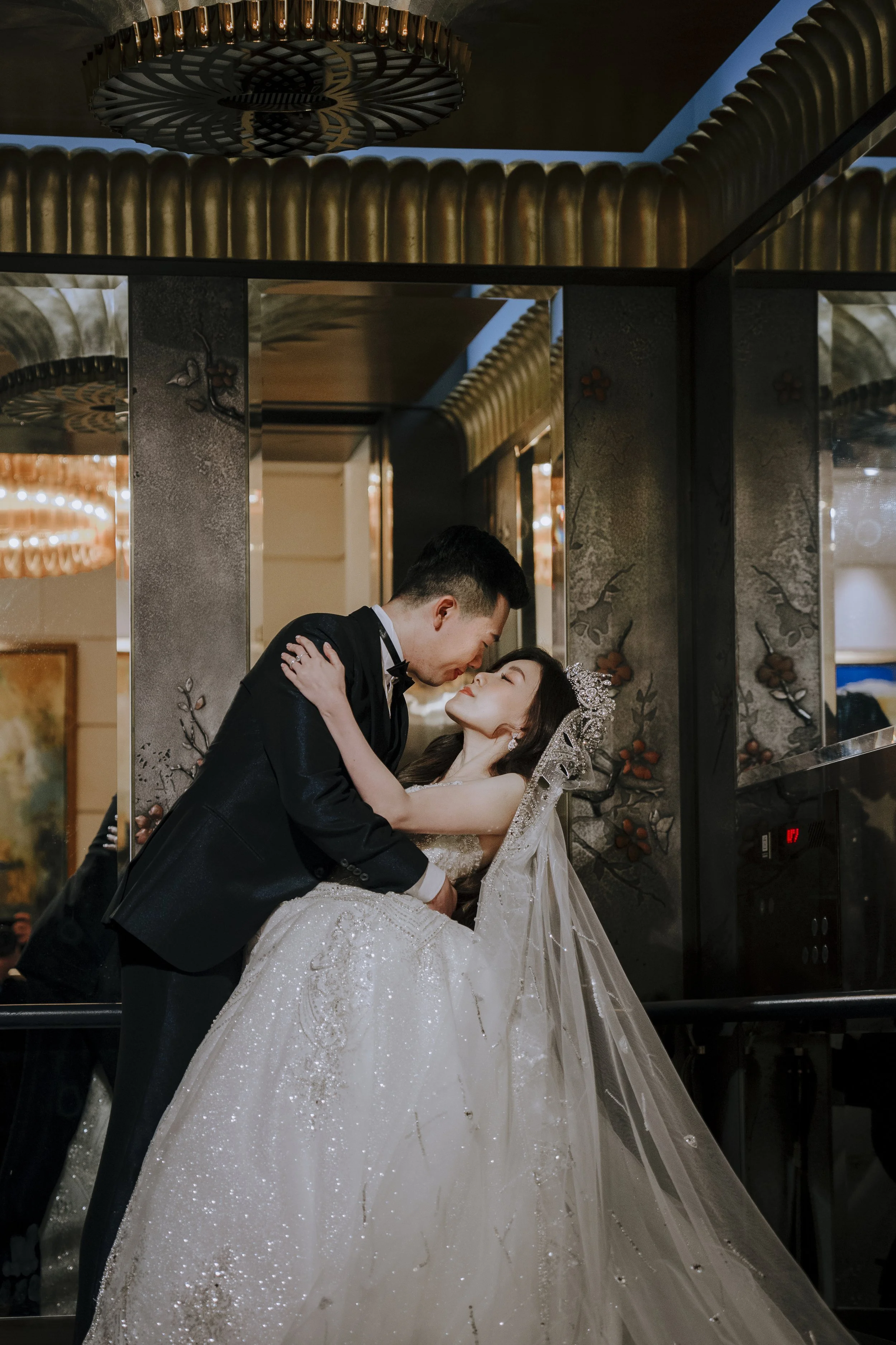 A groom in a tuxedo and a bride in a wedding gown are sharing a romantic moment in an elevator. The groom is holding the bride's waist and leaning towards her, while the bride is gazing up at him. The scene is decorated with ornate decorations on the elevator walls and warm lighting in the background.