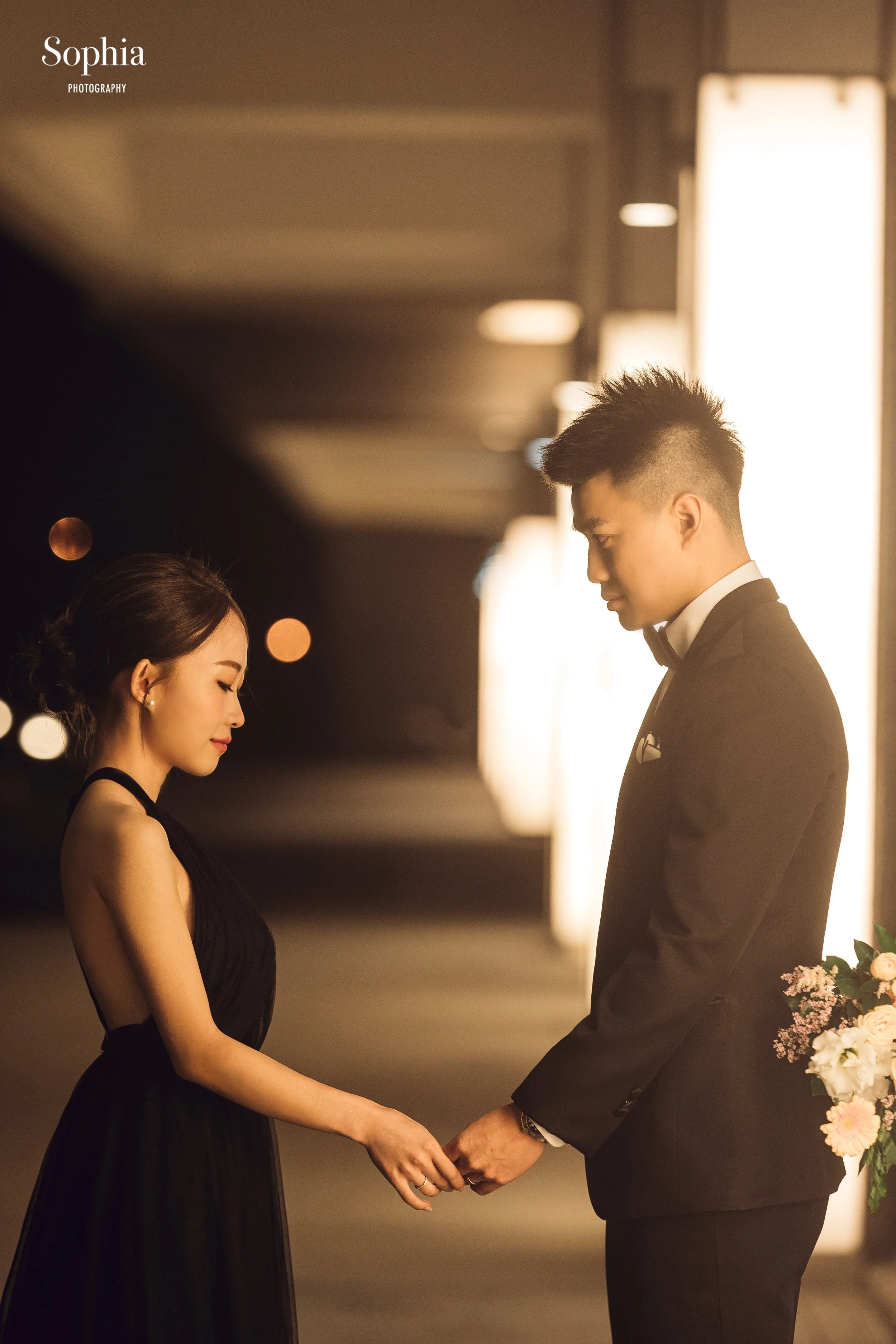 夜間外景婚紗攝影，新人穿著黑色禮服與西裝在暖色光影中牽手