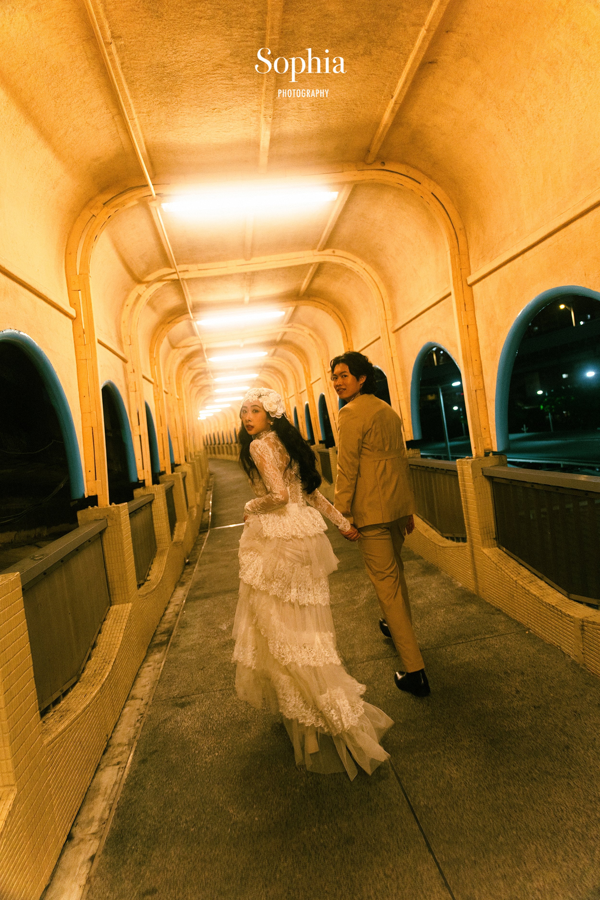 A bride in a lace wedding dress holding hands with a groom in beige suit walking through a yellow corridor at night.
