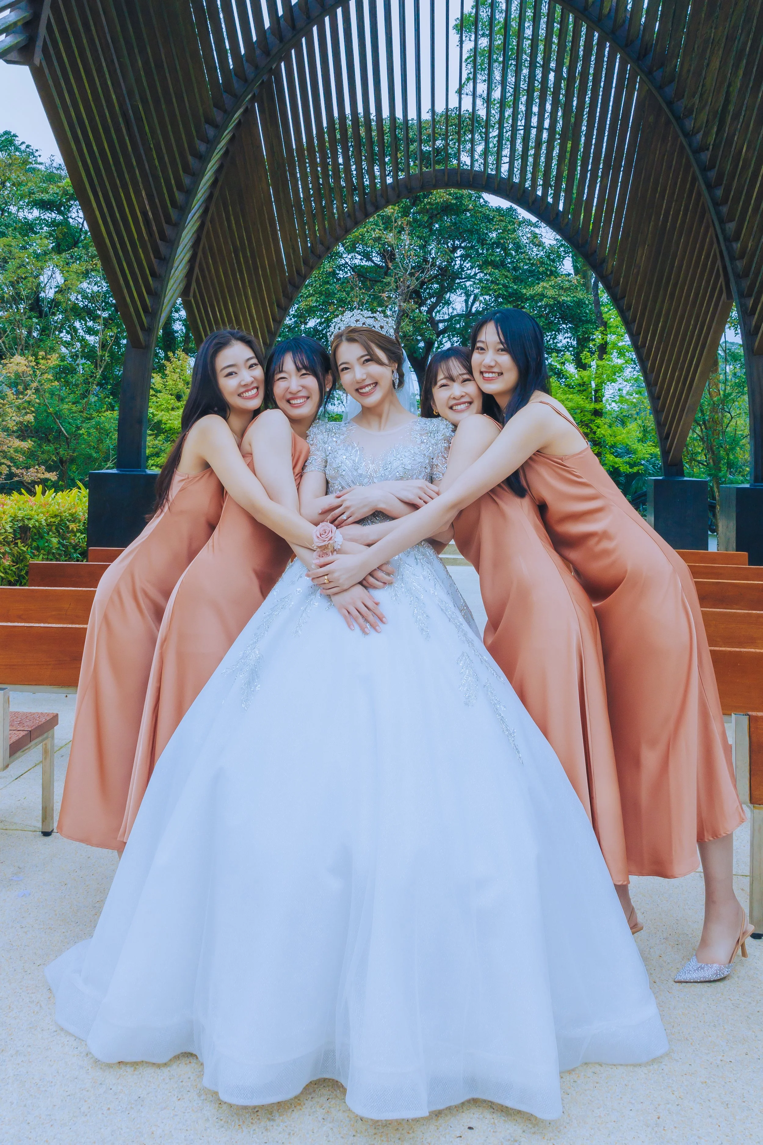 A group of five women, with one woman dressed as a bride in a white wedding gown, and four women dressed in matching peach-colored dresses, hugging and smiling in an outdoor park with a large wooden arch structure and green trees in the background.