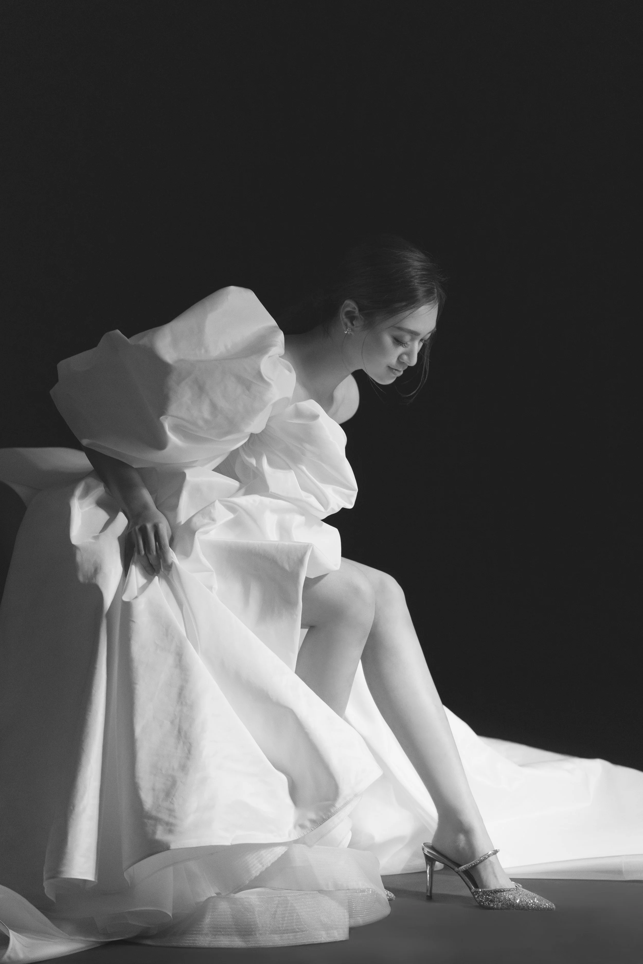 A woman in a white dress with ruffled sleeves is sitting down, looking down. She is wearing high-heeled shoes with glitter.