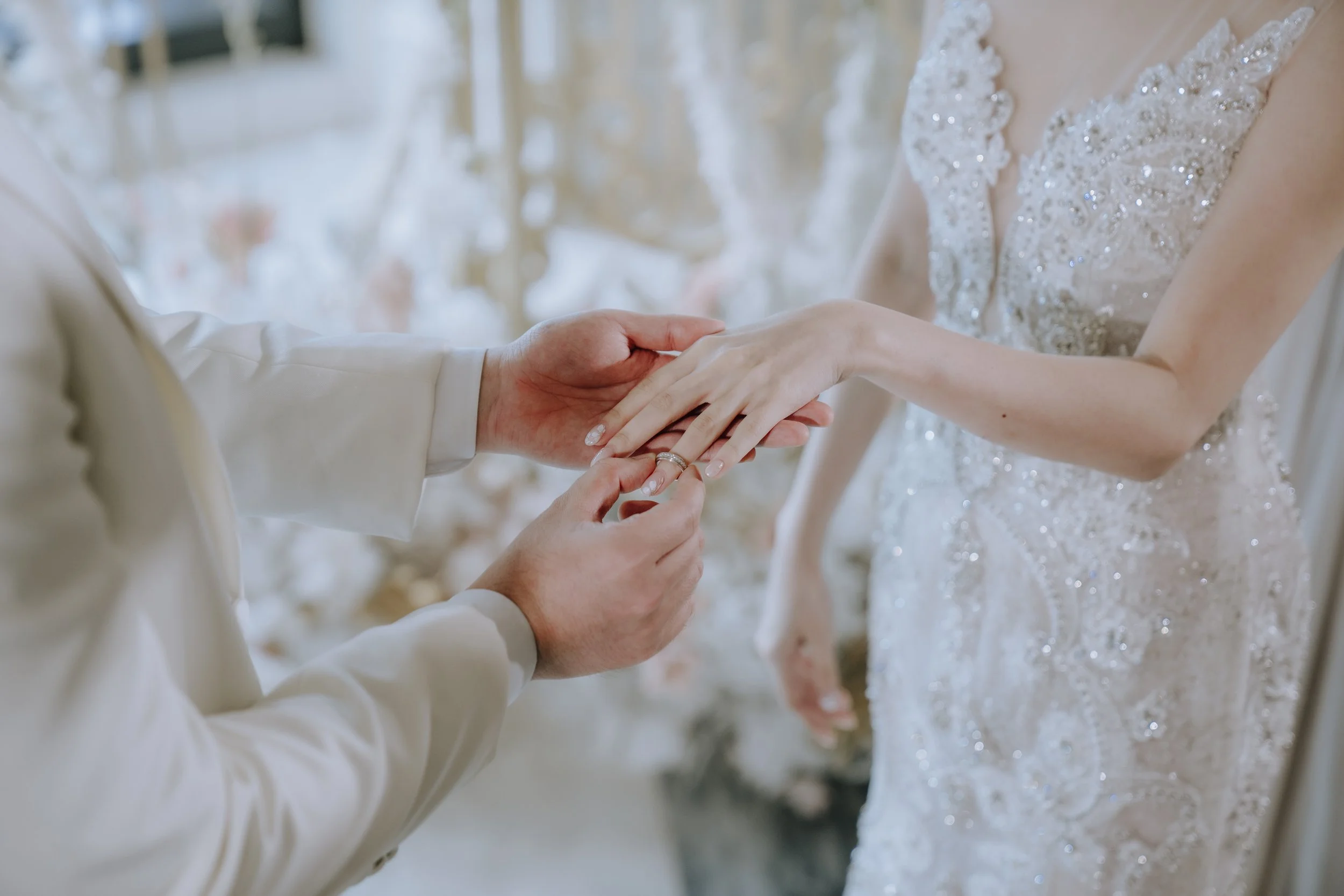 A couple exchanging wedding rings during a wedding ceremony, with the bride wearing a beaded wedding gown and the groom in a light-colored suit.