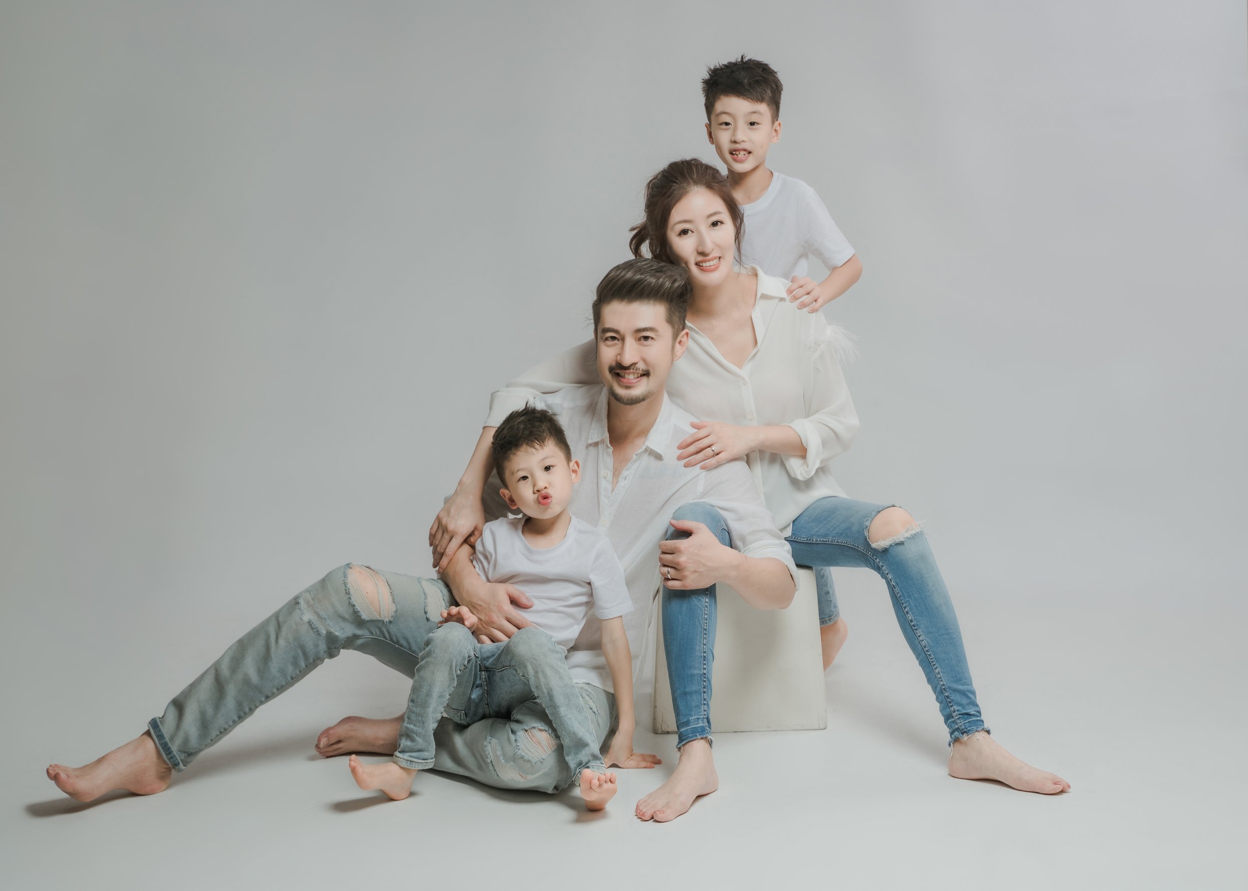 A family of four, including a man, woman, and two boys, posing together in a studio with a plain light gray background. They are all smiling and wearing casual light-colored clothing.