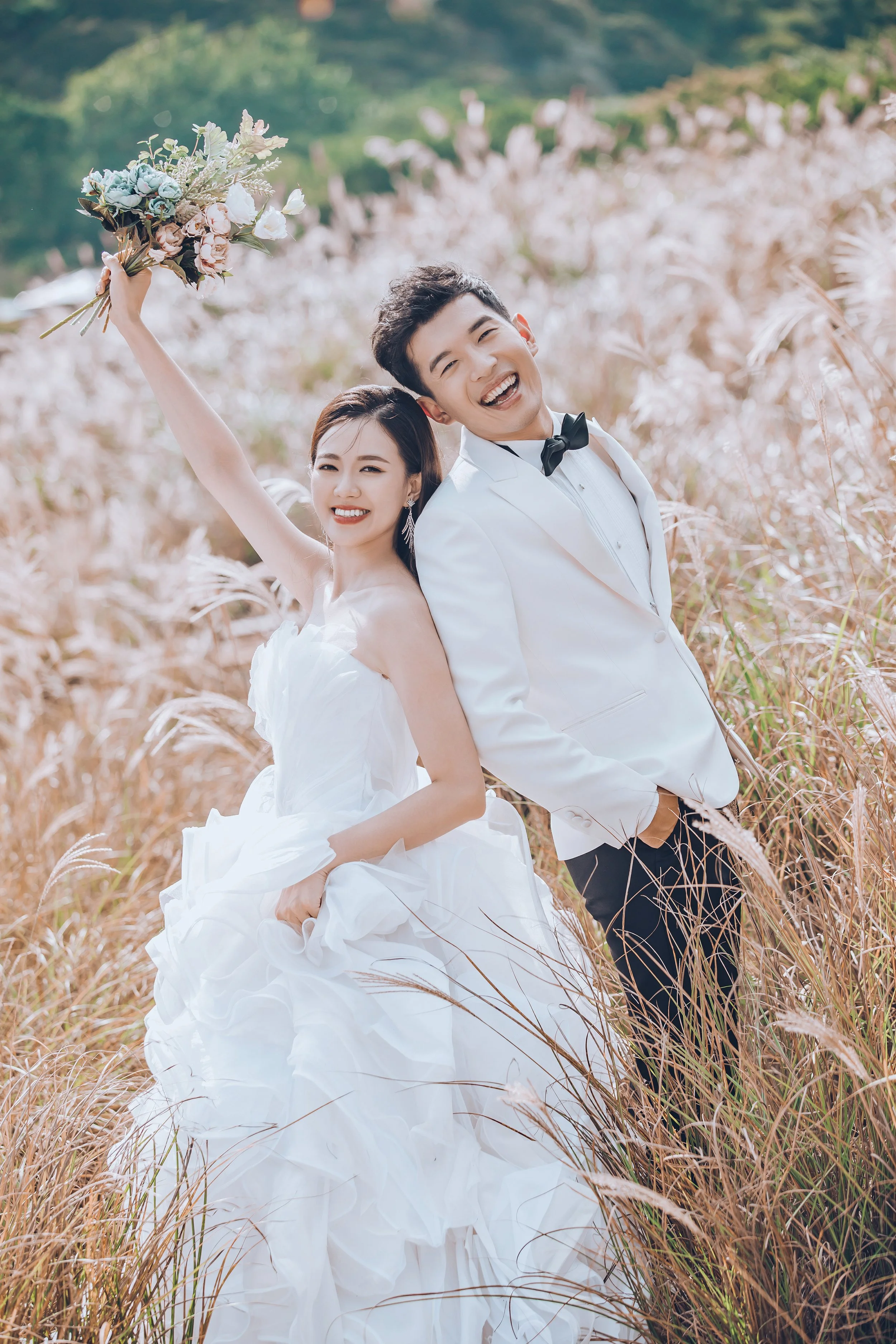 A happy couple in wedding attire standing in a field of tall, beige grass with a blurred green background. The bride is holding a bouquet of flowers and has a joyful expression, while the groom is laughing, dressed in a white suit with a black bow tie.