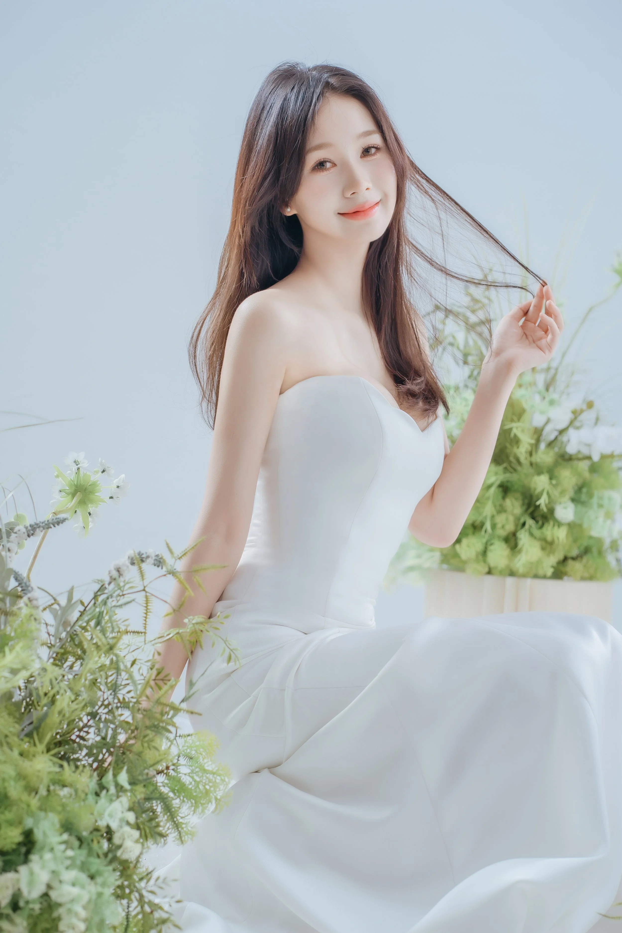 A woman with long brown hair wearing a white strapless dress sitting among green plants and flowers, smiling gently.