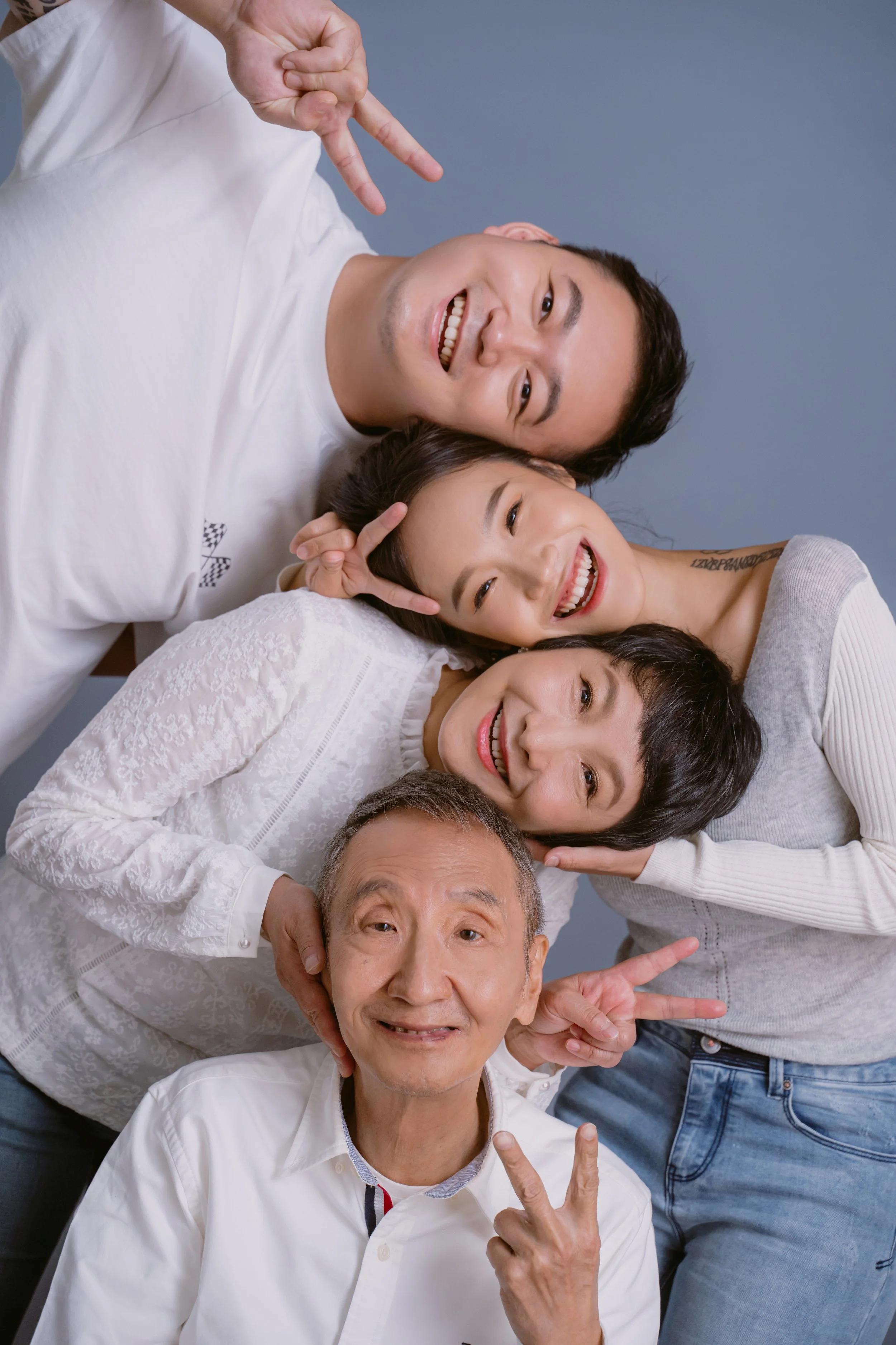 Group of five people lying on the floor, smiling and making peace signs with their hands, close together, against a blue background.