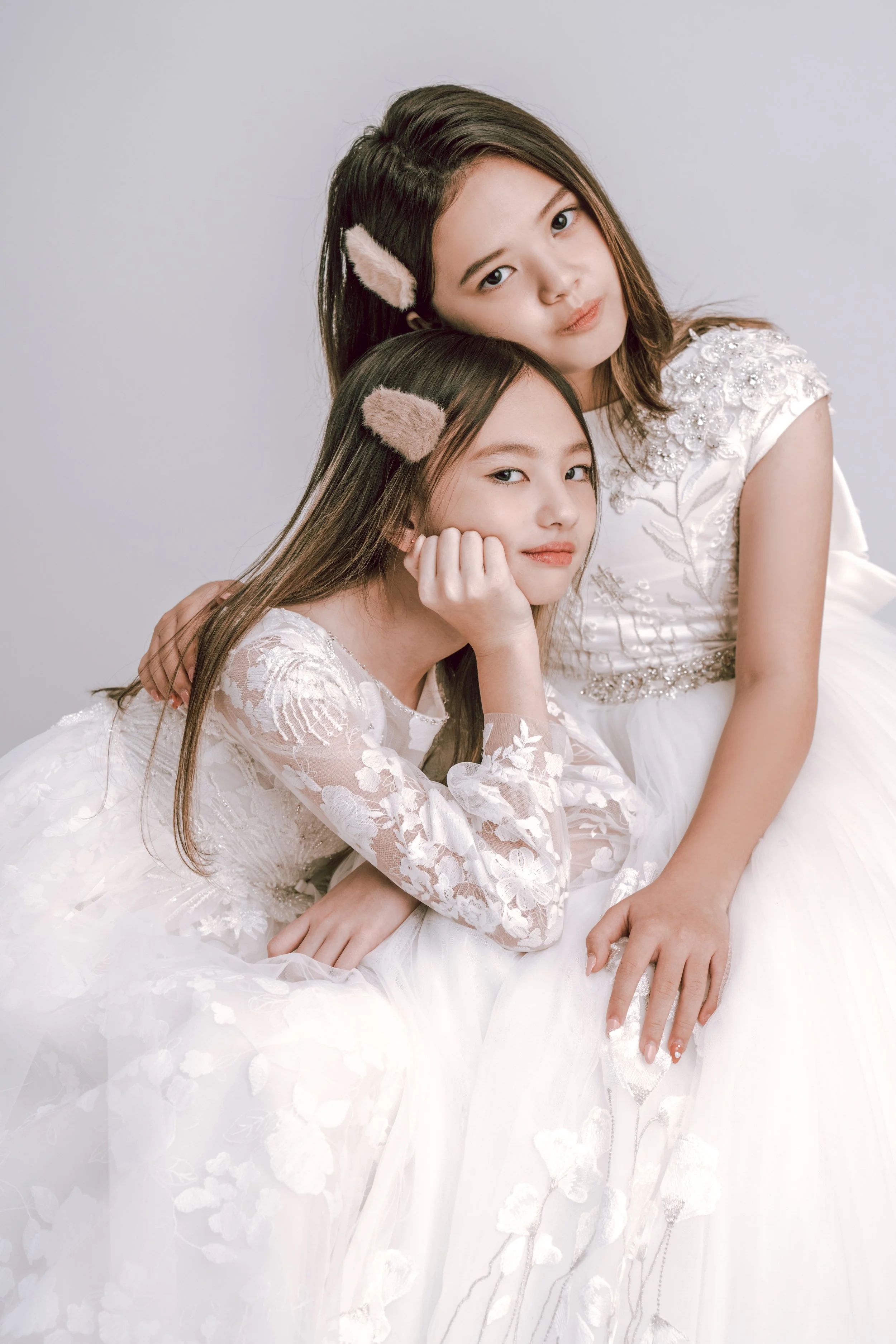 Two young girls with long hair wearing white dresses and furry hair accessories, sitting close together against a plain light background.